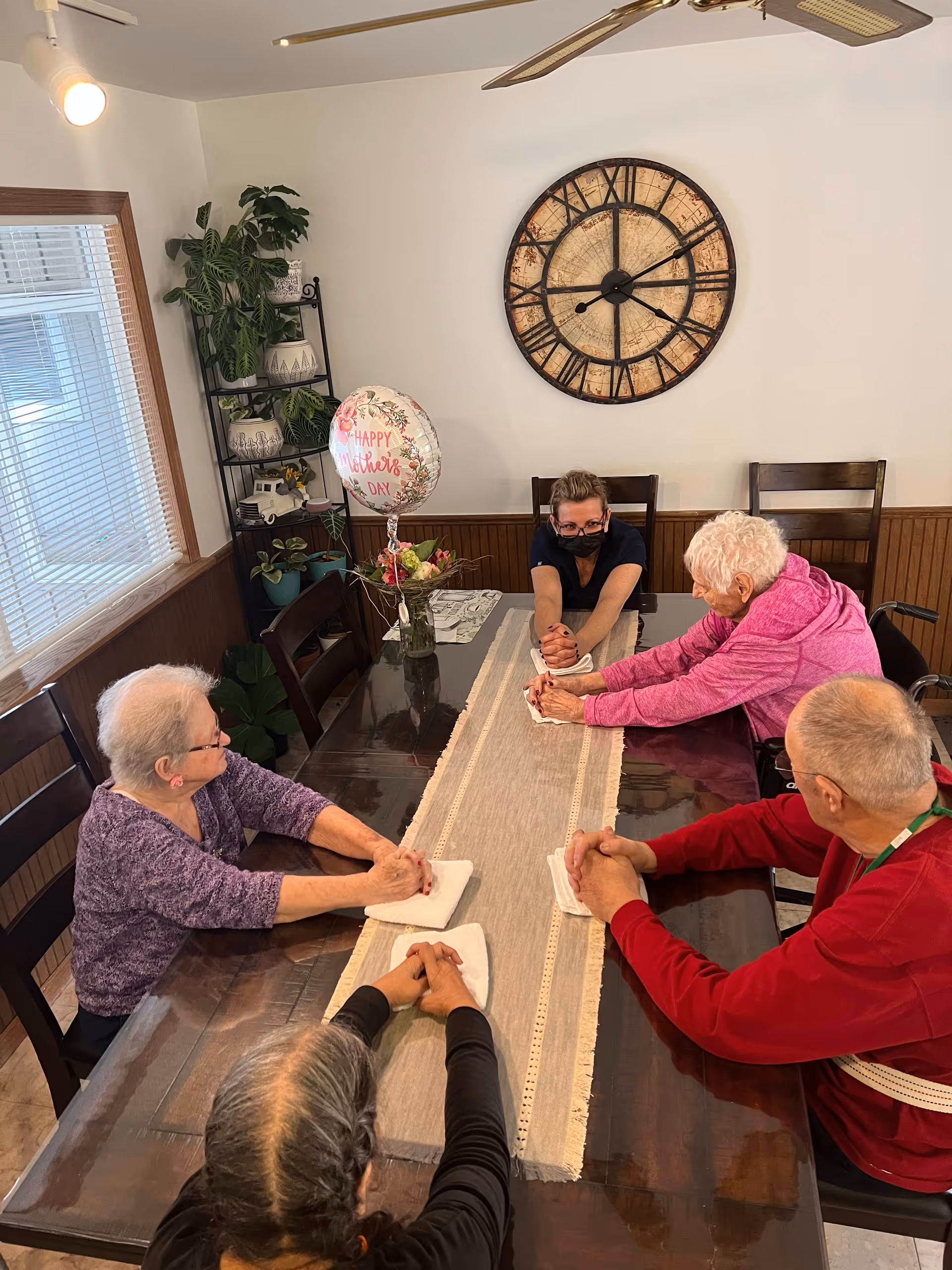 A group of older adults and a masked caregiver seated around a dining table holding hands, with a 'Happy Mother's Day' balloon and a large wall clock behind them.