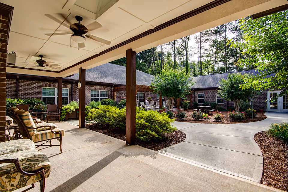 Covered outdoor patio area with ceiling fans and cushioned chairs overlooking a landscaped courtyard with bushes, trees, and a paved walkway surrounded by a brick building.