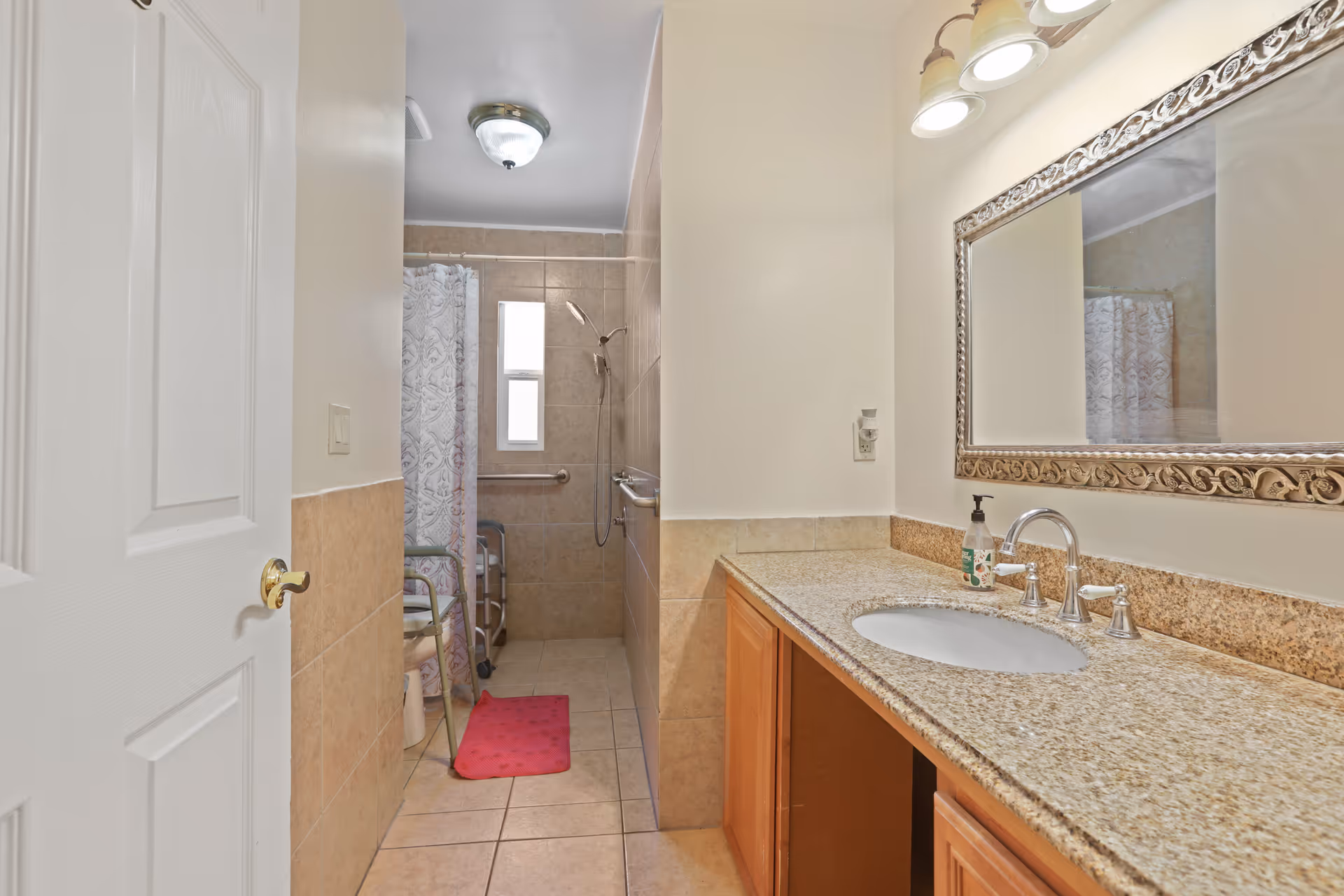 A bathroom with beige tiled floor and walls, featuring a granite countertop with a sink and a large ornate mirror above it. There is a shower area with a white patterned shower curtain, a grab bar, and a shower chair. A red bath mat is placed on the floor near the shower.