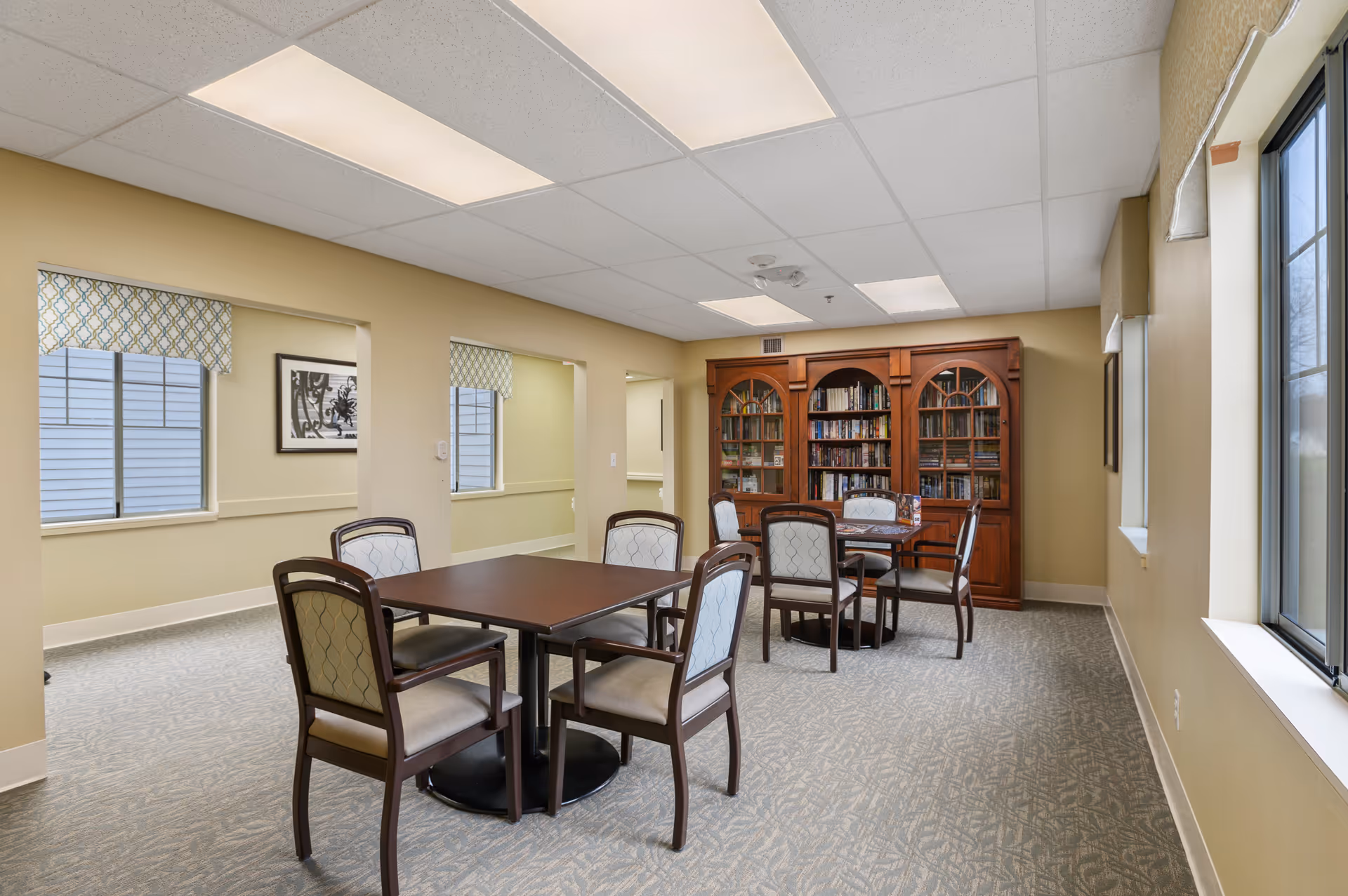 A well-lit senior living lounge with tables, chairs, and a wooden bookcase.