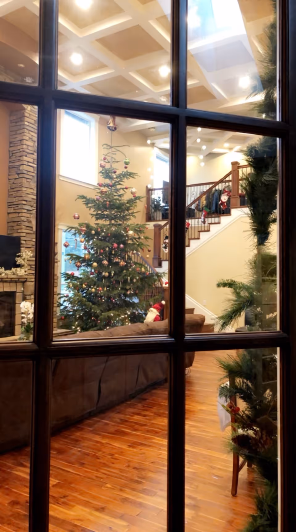 Interior living room seen through window panes featuring a decorated Christmas tree, staircase, sofa, fireplace, and hardwood floors.