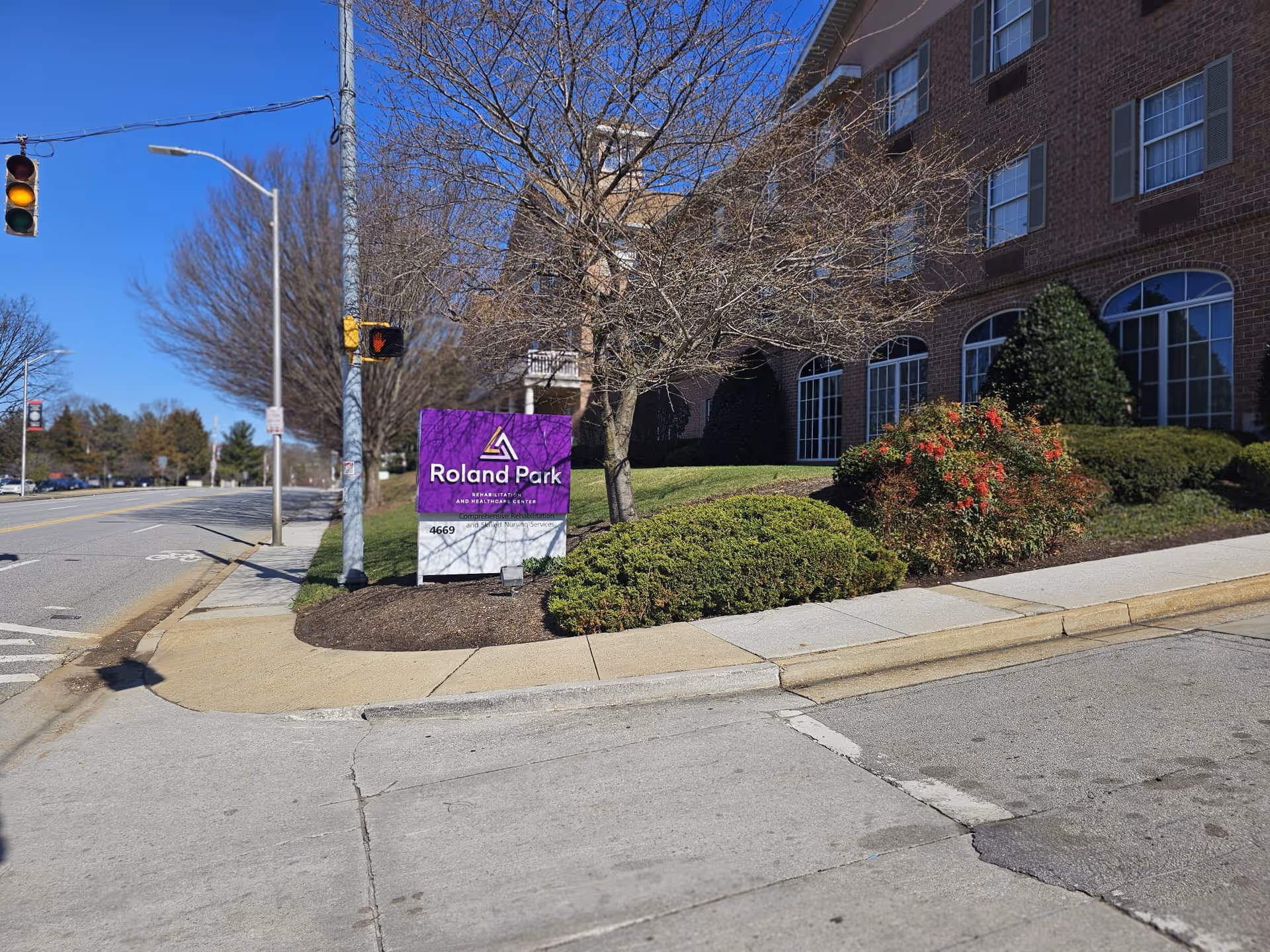 Exterior view of Roland Park Rehabilitation and Healthcare Center showing a brick building with multiple windows, a sidewalk, landscaped bushes and trees, and a purple sign with the facility's name near a street intersection with a traffic light.