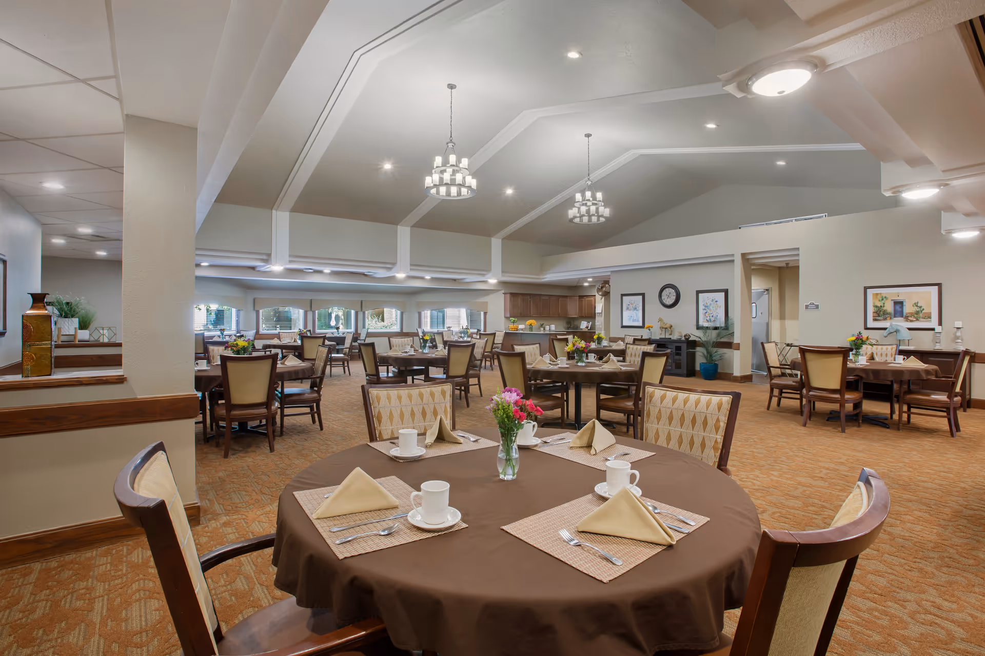 A spacious dining room in a senior living facility with multiple round tables covered with brown tablecloths, each set with beige napkins, white cups, and silverware. The room features high ceilings with modern chandeliers, beige walls, carpeted floors, and large windows letting in natural light. There are decorative plants and framed artwork on the walls.