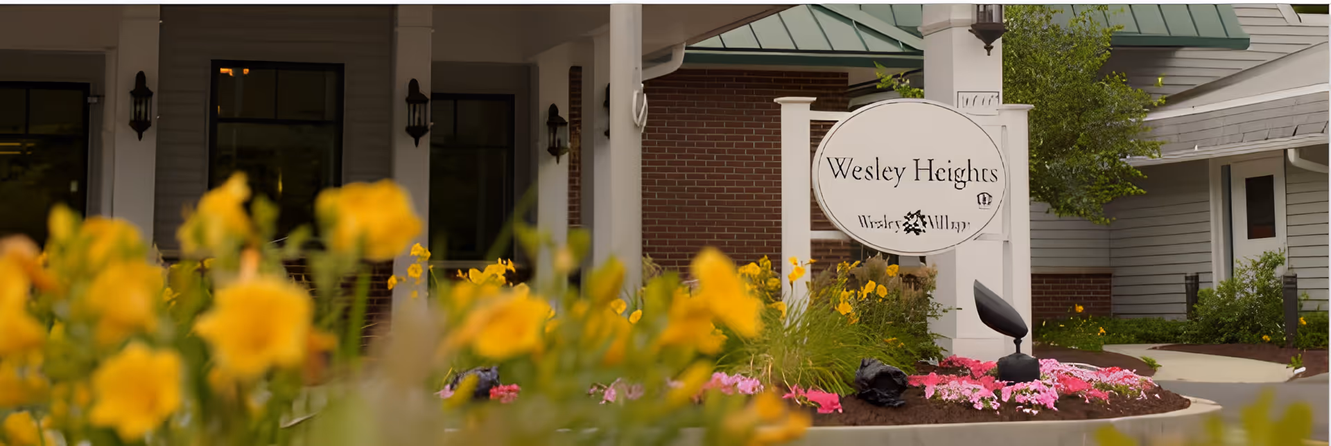 Entrance area of Masonicare at Wesley Heights facility with a white sign displaying 'Wesley Heights' and surrounding colorful flowers including yellow and pink blooms, with part of the building and greenery visible in the background.
