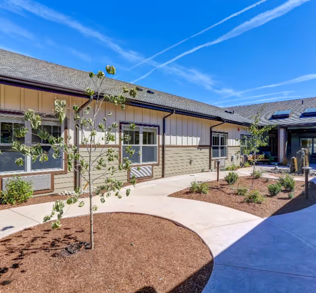 Outdoor courtyard area of a senior living facility with a paved walkway, young trees, and small landscaped garden beds under a clear blue sky.