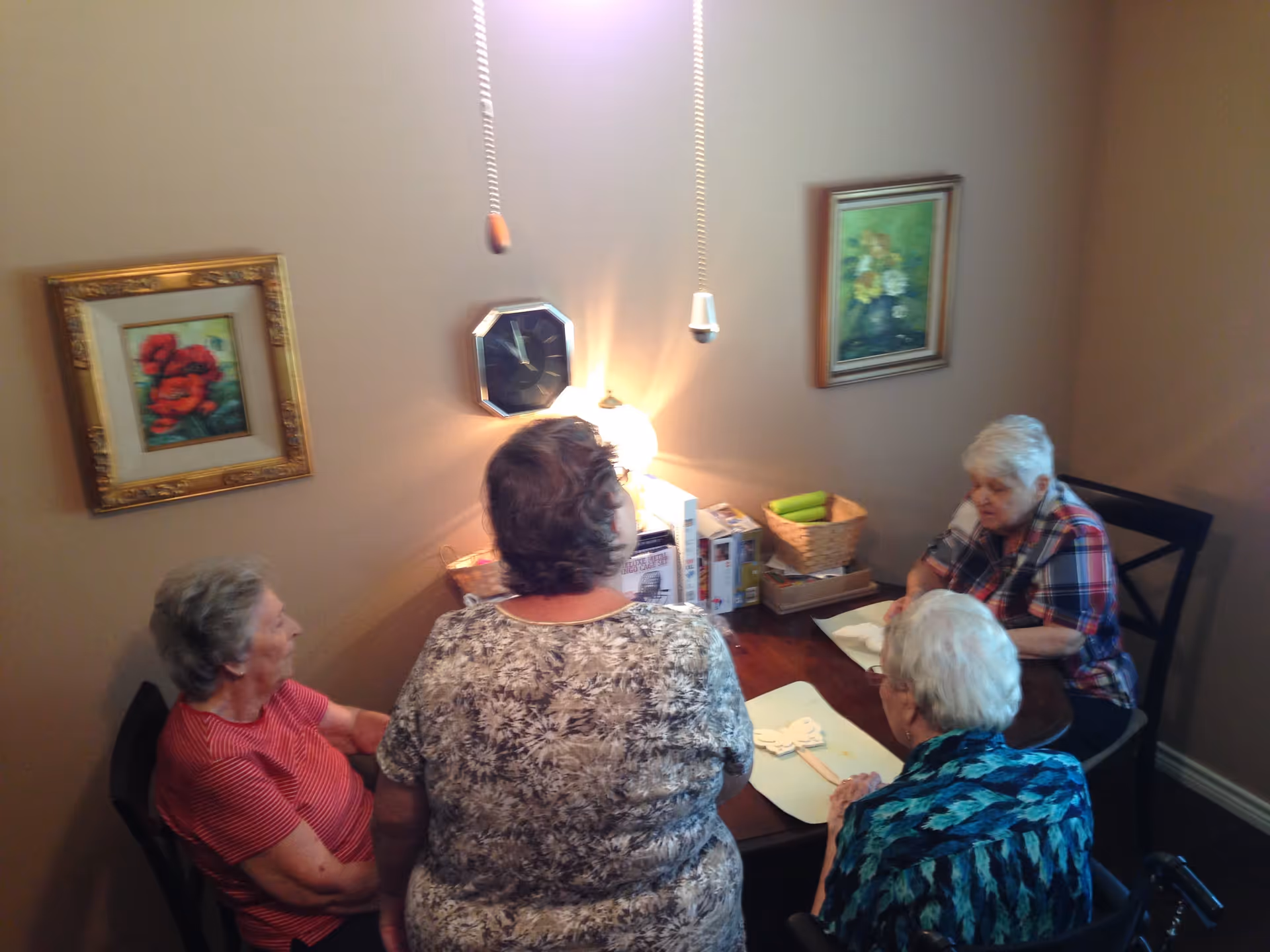 Four elderly women sitting around a wooden dining table in a room with beige walls. Two framed floral paintings hang on the walls, and a clock is mounted between them. A small lamp on the table provides light, and there are some boxes and a basket with green towels on the table.