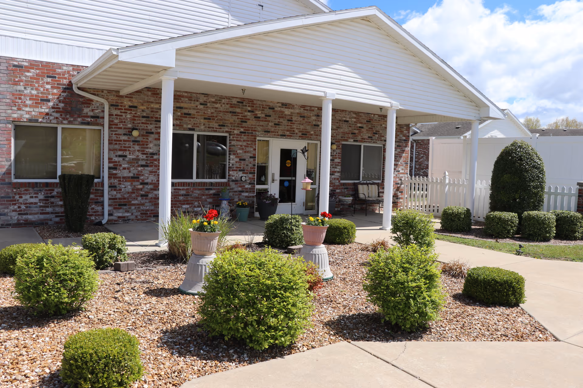 Front entrance of a brick building with a white covered porch supported by white columns. The area in front has neatly trimmed bushes, flower pots with colorful flowers, and a paved walkway. There is a white picket fence and a clear blue sky with some clouds in the background.