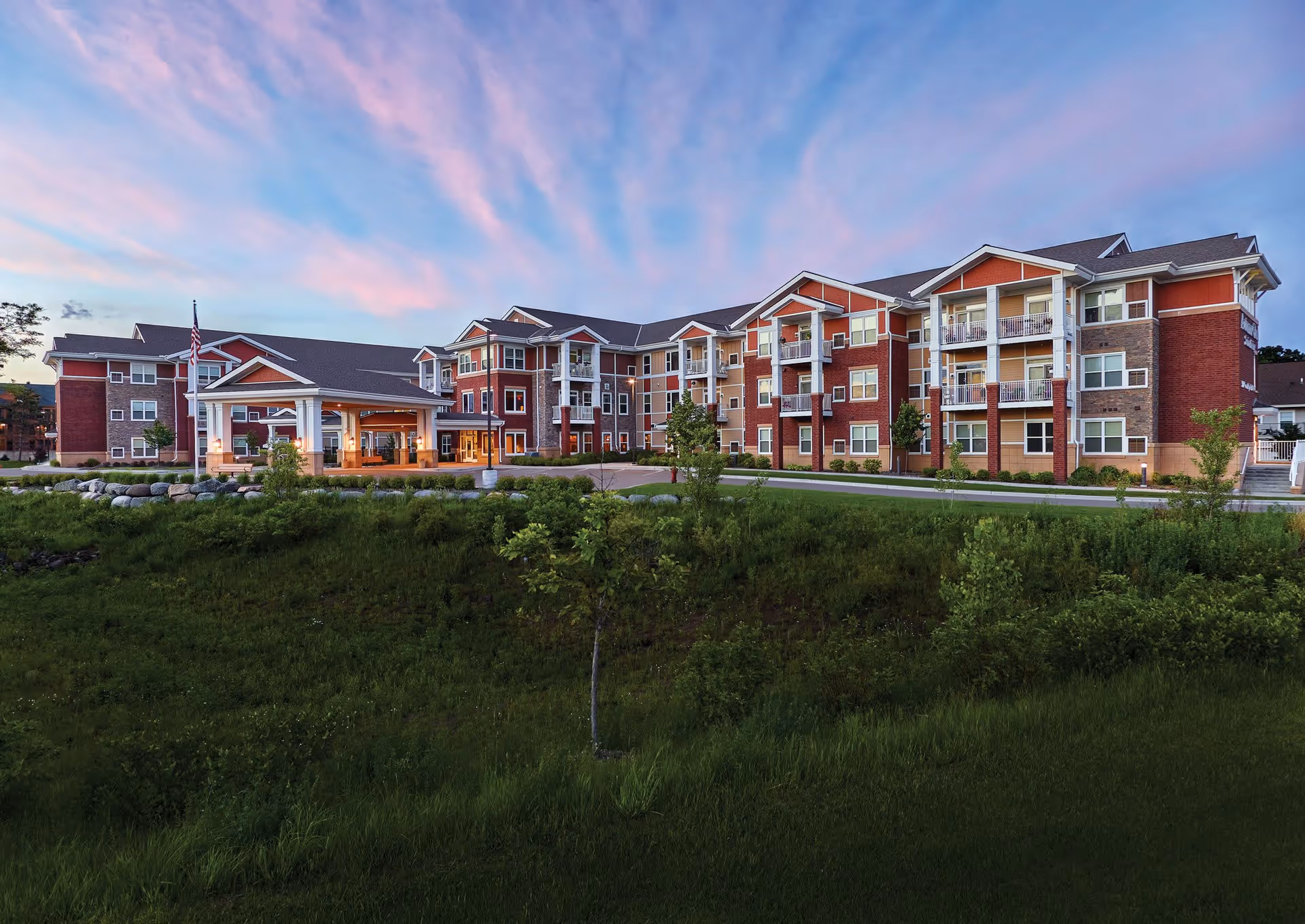 Exterior view of Autumn Glen Senior Living facility during sunset with a large multi-story building featuring balconies, a covered entrance, and well-maintained landscaping with green grass and small trees in the foreground.
