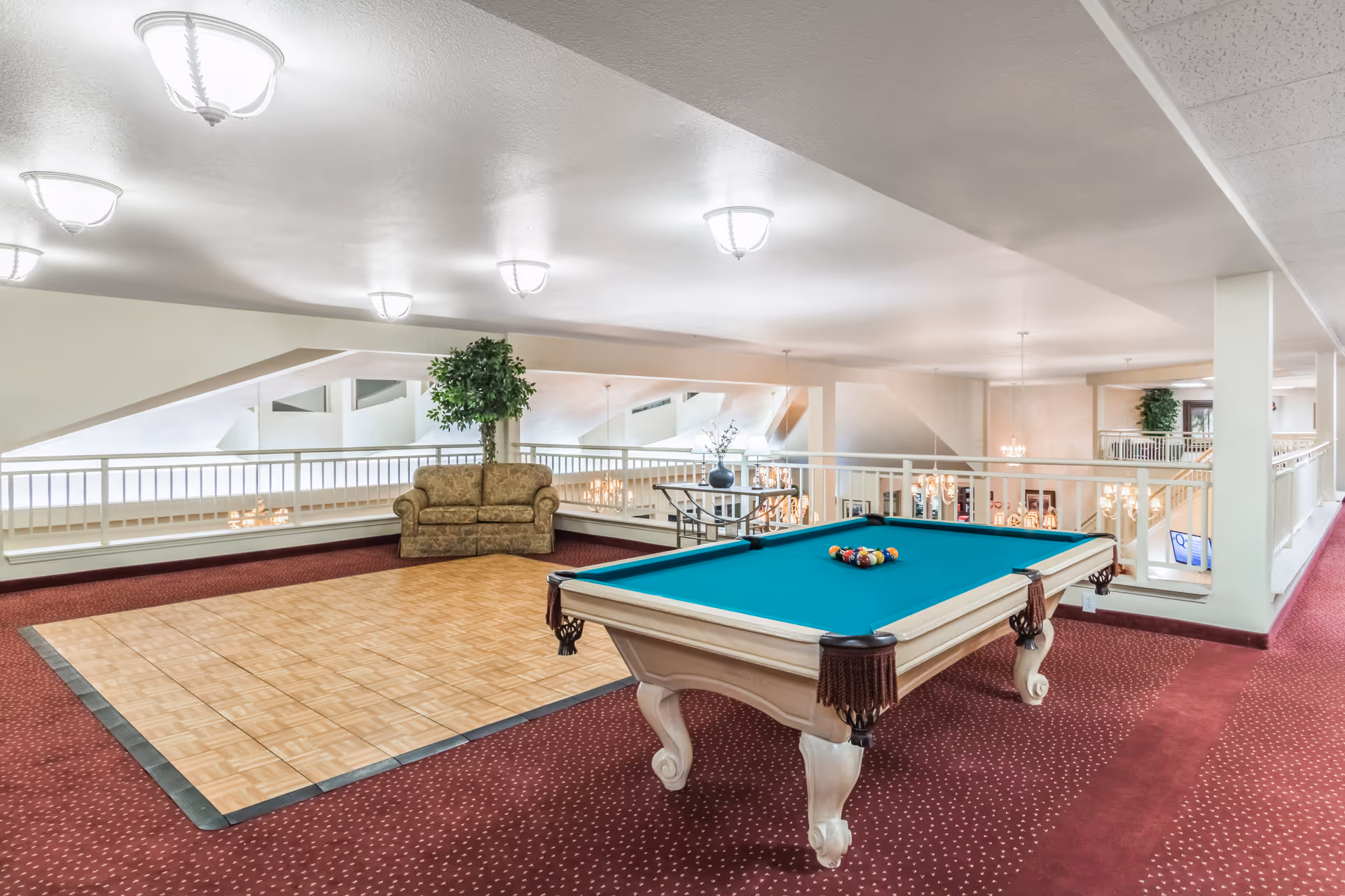 Interior view of a senior living facility common area featuring a pool table with balls arranged for a game, a small wooden dance floor, a patterned sofa, a potted plant, and a railing overlooking a lower level with chandeliers and additional seating.