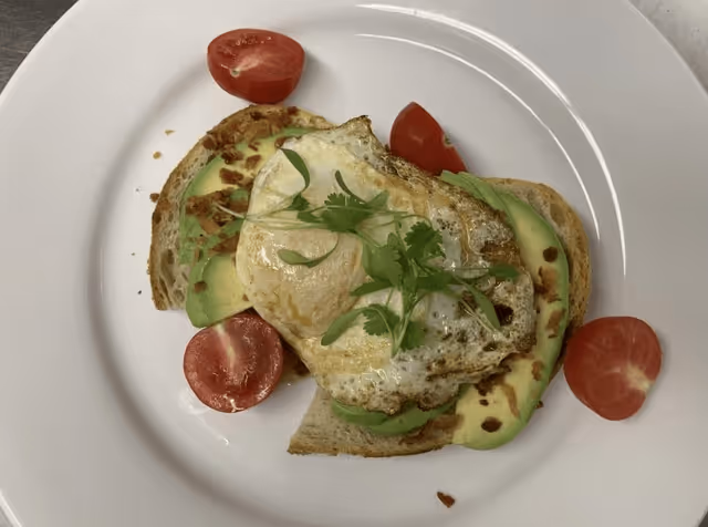 A plate with a slice of toasted bread topped with sliced avocado, a fried egg, and garnished with fresh herbs, surrounded by halved cherry tomatoes.