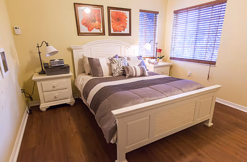 A cozy bedroom with a white wooden bed frame and a neatly made bed featuring multiple pillows and a striped comforter. There are two framed floral artworks hanging above the headboard. On either side of the bed are white nightstands, one with a lamp and a printer, and the other with a lamp and a small plant. The room has wooden flooring and two windows with wooden blinds allowing natural light to enter.