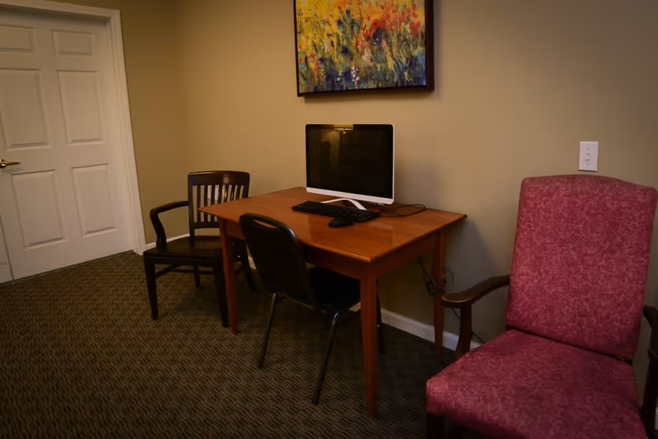 A small room with a wooden table holding a computer monitor, keyboard, and mouse. There are three chairs around the table: a black chair, a wooden chair, and a pink upholstered armchair. A colorful abstract painting hangs on the beige wall above the table, and a closed white door is visible on the left side.