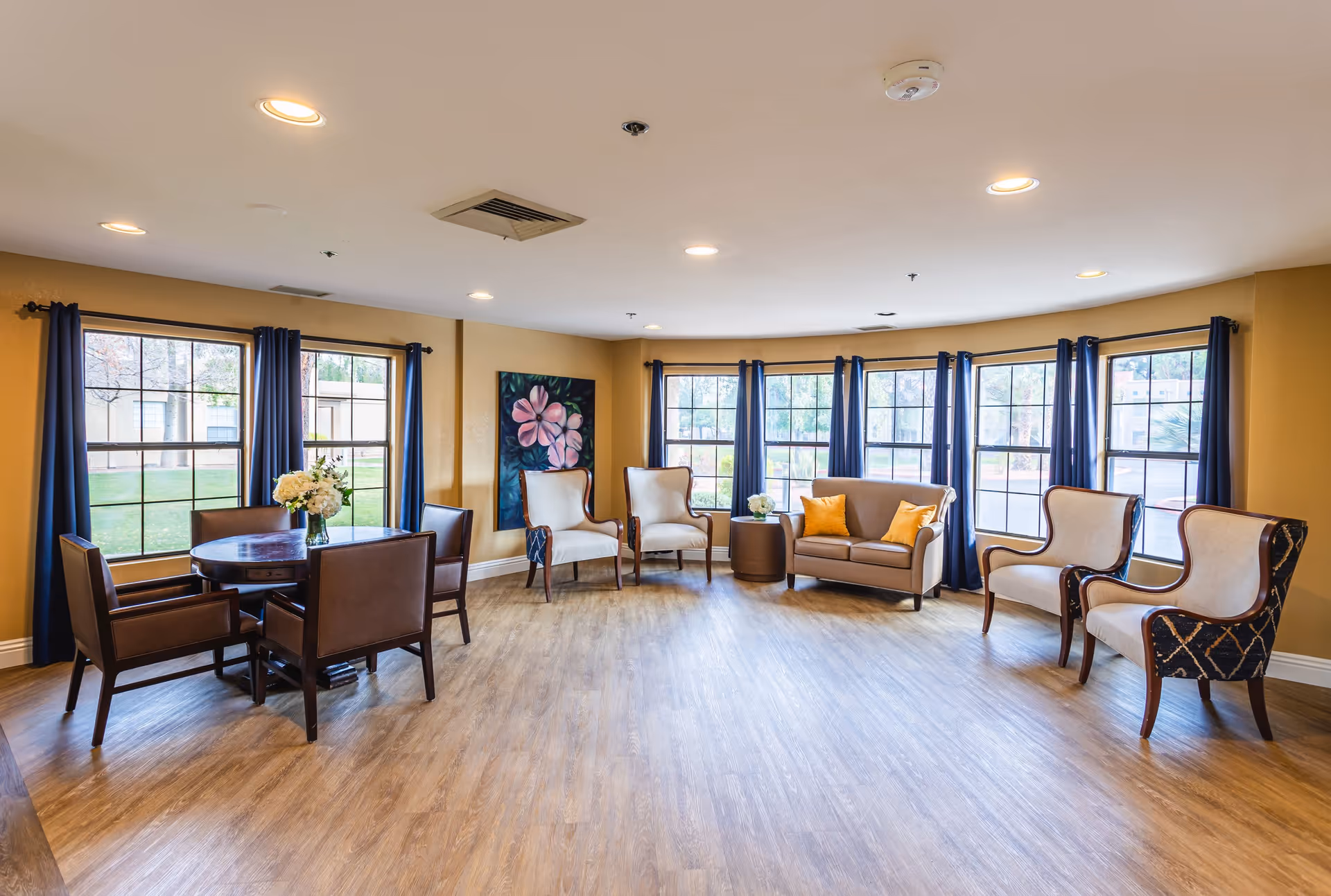 A bright and spacious living room area with large windows covered by dark blue curtains. The room features a round wooden table with four brown leather chairs, a small sofa with two yellow pillows, and four upholstered armchairs arranged around the room. The walls are painted a warm beige color and decorated with a floral painting. The floor is a light wood laminate.