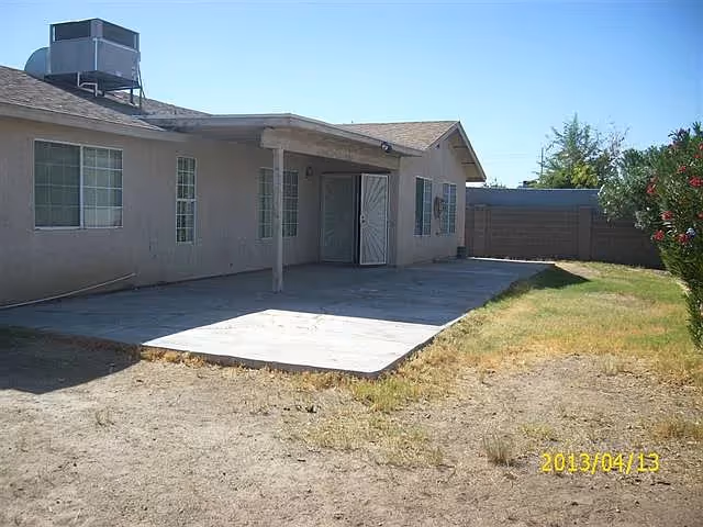 Rear exterior of a single-story building showing a covered patio, concrete slab, and a sparse grassy yard.