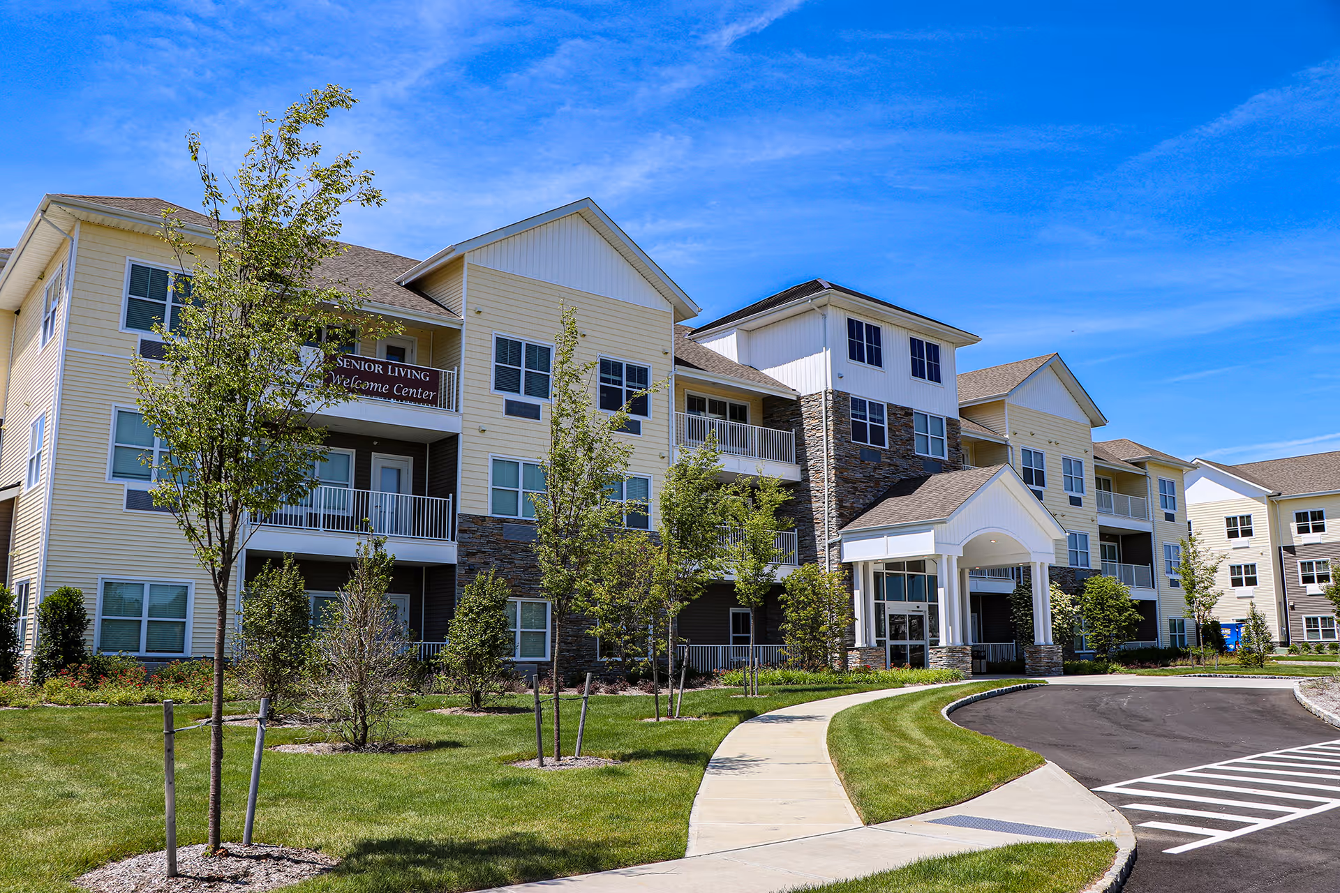 Exterior view of a senior living facility building with multiple floors, balconies, and a covered entrance. The building is surrounded by a well-maintained lawn, young trees, and a curved driveway under a clear blue sky.