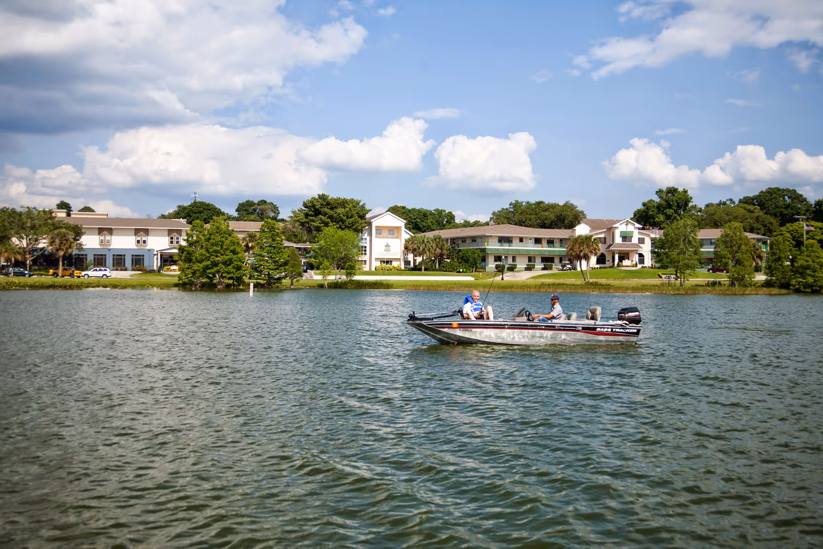 Two people fishing on a boat in a lake with a senior living facility building and trees in the background under a partly cloudy sky.
