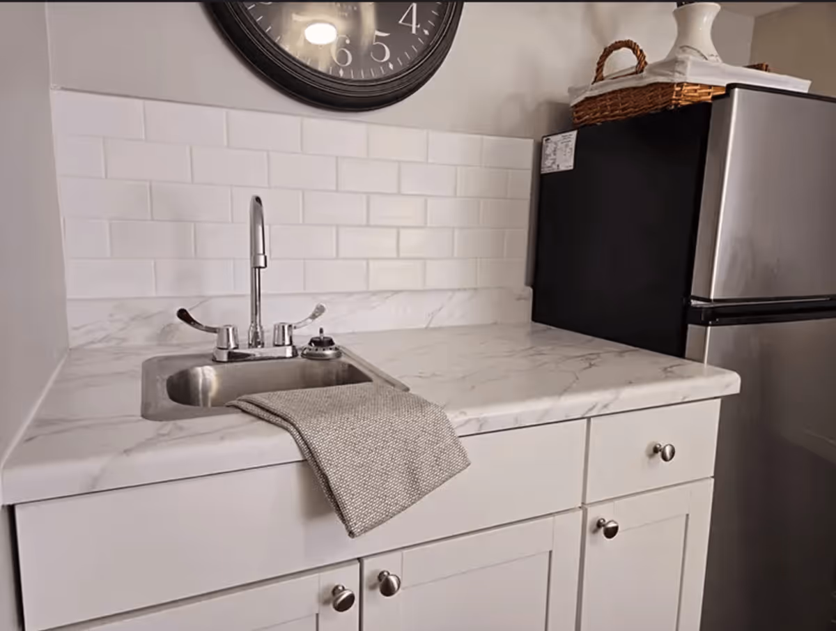 A small kitchen area with a stainless steel sink, a gray dish towel draped over the edge, white cabinets with silver knobs, a white marble countertop, white subway tile backsplash, a black and silver mini refrigerator, and a large black wall clock above the sink.