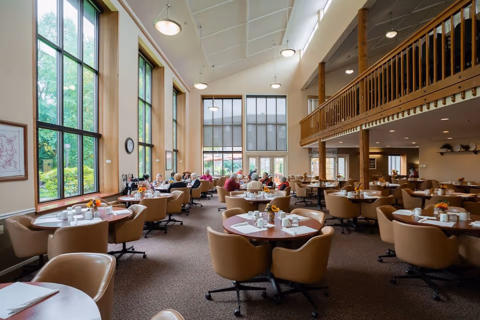 A spacious dining room with large windows letting in natural light. Several round tables with beige chairs are set with white cups, plates, and napkins. A group of elderly people are seated at tables near the windows, engaged in conversation. The room features high ceilings with hanging lights and a wooden balcony railing on the upper level.