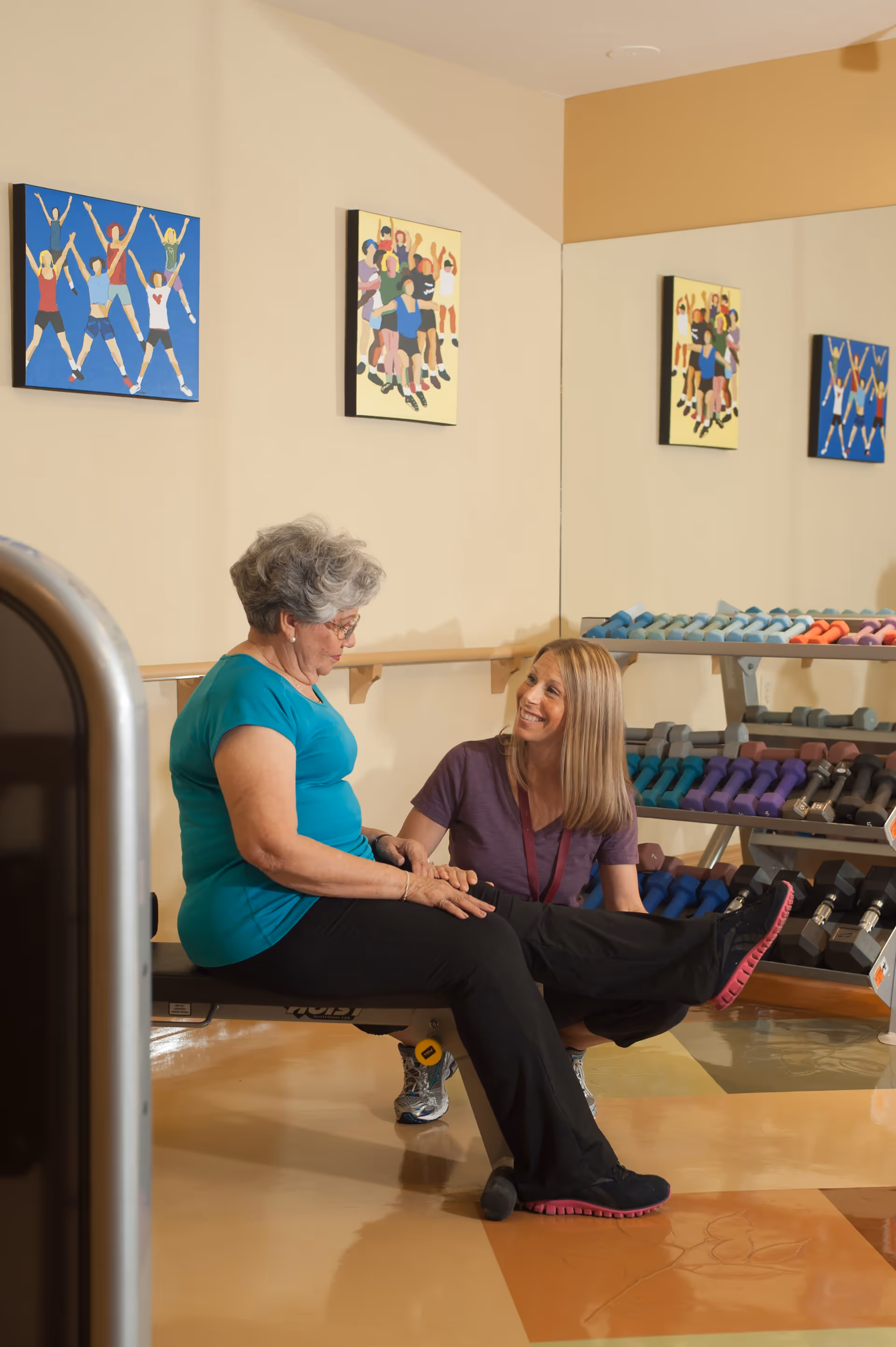 An elderly woman in a turquoise shirt and black pants is seated on a workout bench lifting one leg, while a younger woman with blonde hair in a purple shirt kneels beside her, smiling and offering support. Behind them is a rack filled with colorful dumbbells and two paintings of people exercising on the beige wall.