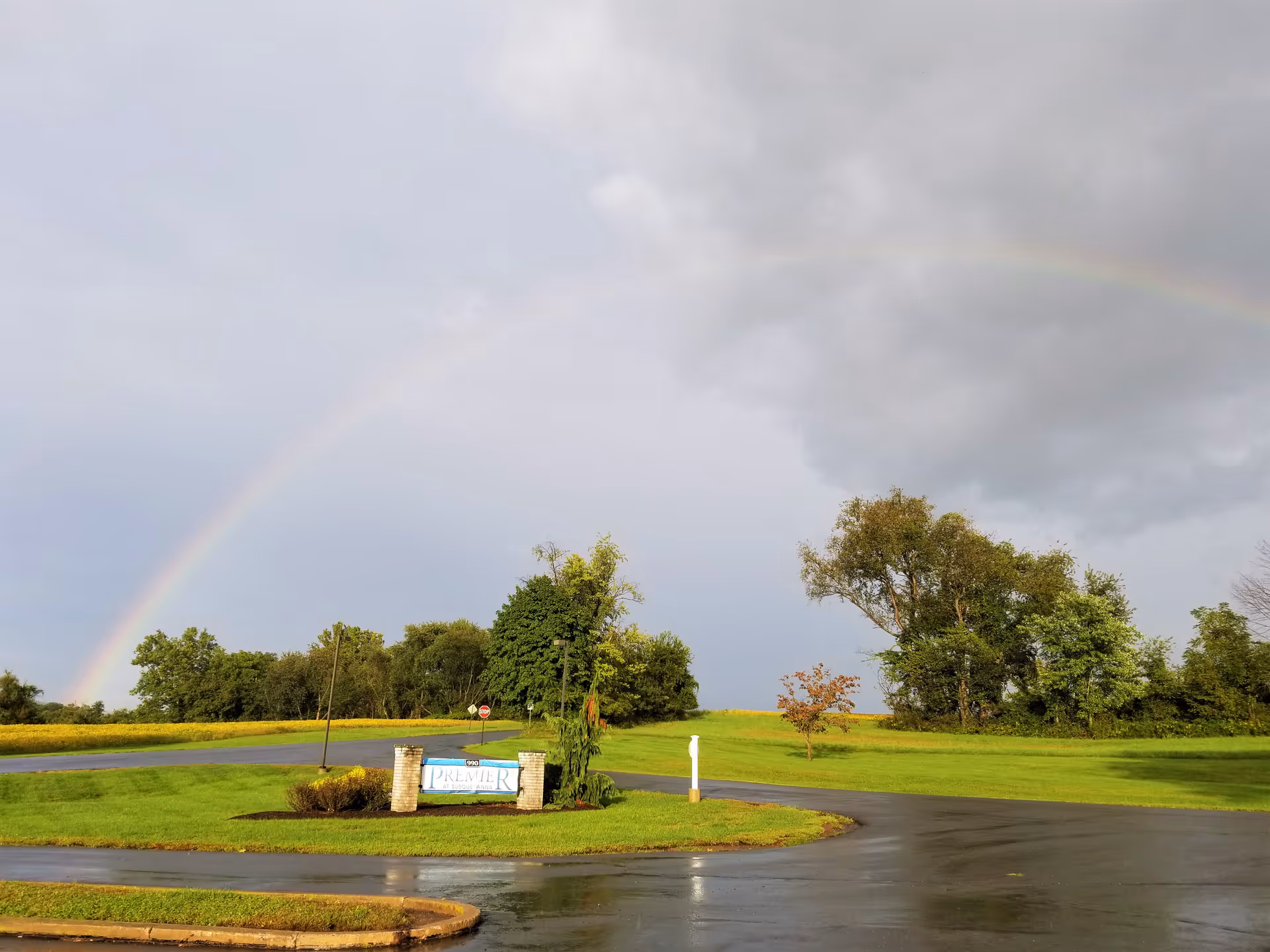 A scenic outdoor view showing a wet paved road curving around a grassy area with a sign that reads 'Premier'. Trees and a cloudy sky with a faint rainbow are visible in the background.