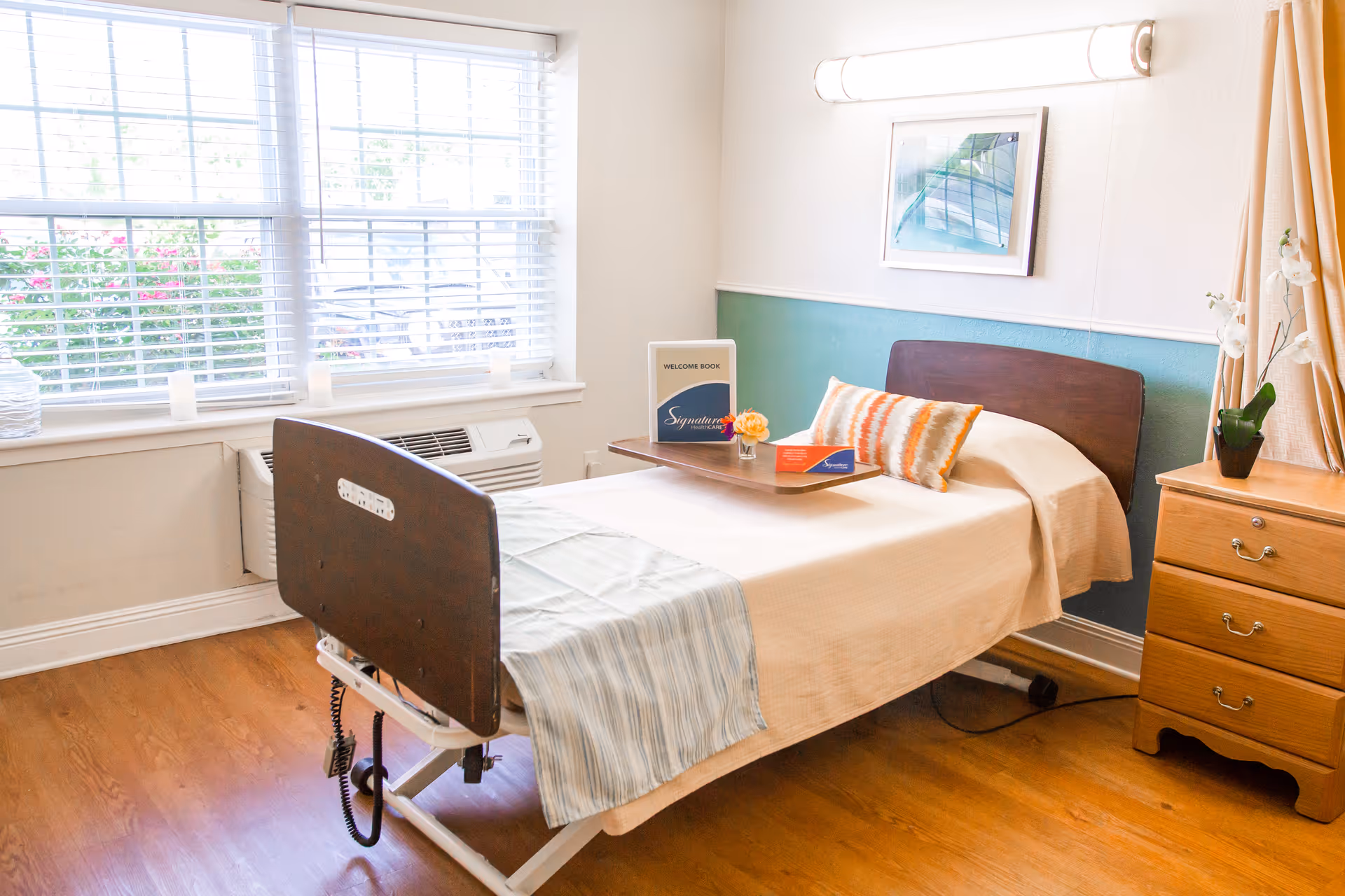 A bright and clean bedroom in a senior living facility with a single hospital-style bed covered with a beige blanket and a striped pillow. A wooden bedside table with three drawers and a potted orchid plant is next to the bed. A tray on the bed holds a welcome book and a small flower arrangement. A large window with white blinds lets in natural light, and a framed picture hangs on the wall above the bed.