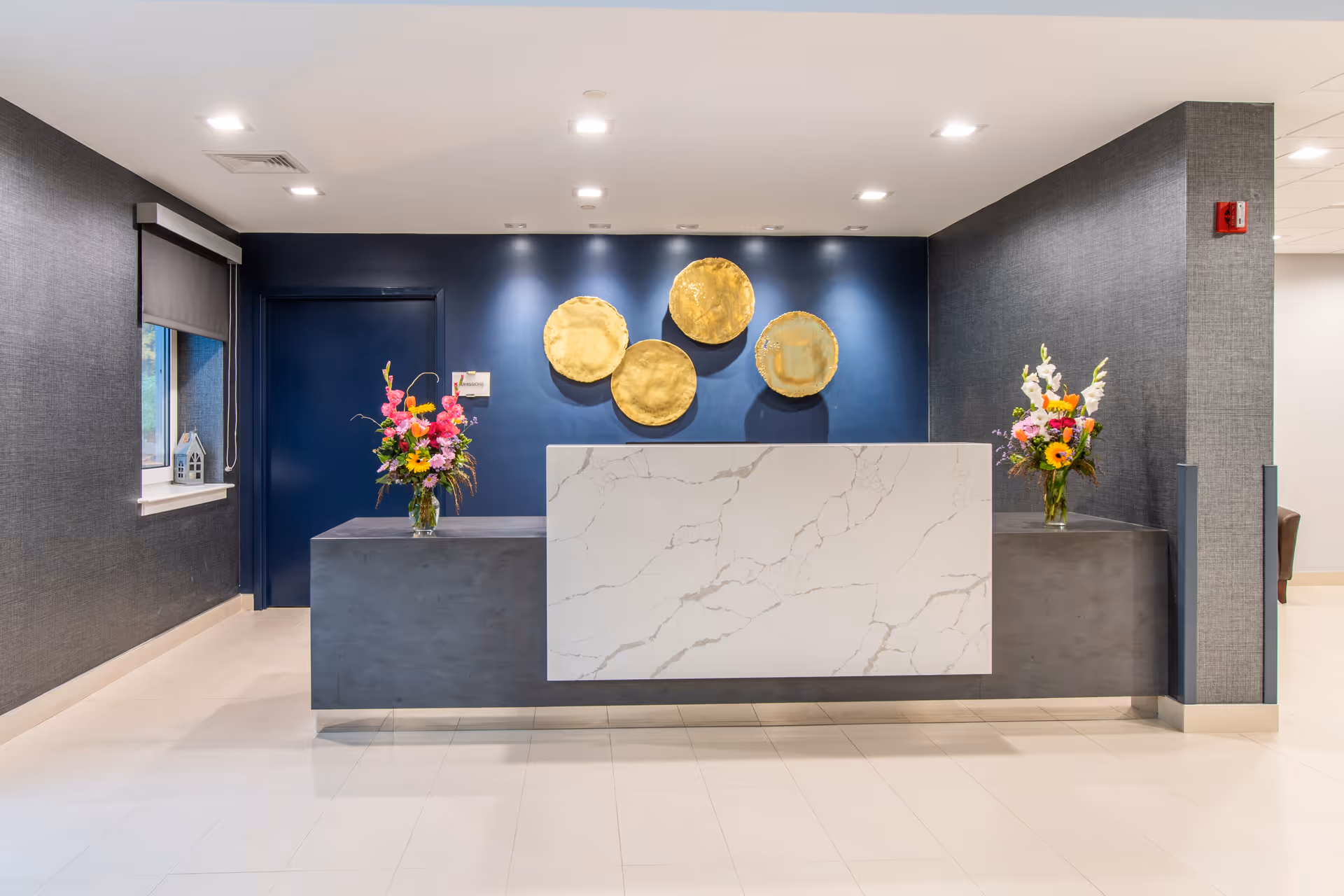 Reception desk area with a marble front panel and two vases of colorful flowers on either side. Behind the desk is a dark blue wall with four round gold decorative wall pieces. To the left is a window with a small decorative house on the sill, and to the right is a gray textured wall with a fire alarm.