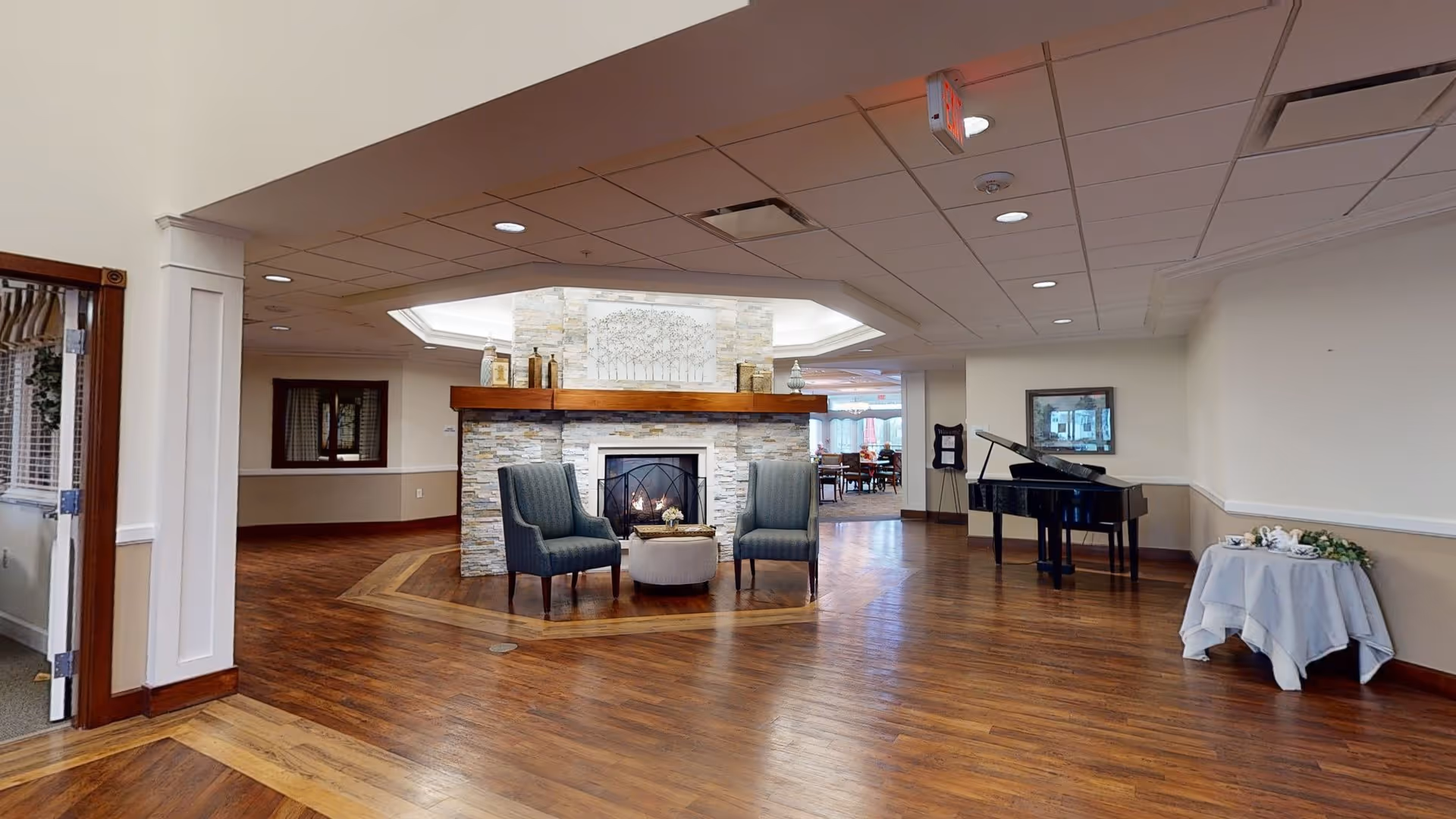 A spacious senior living facility common area with wooden flooring, a stone fireplace with a wooden mantle, two upholstered chairs and a small round ottoman in front of the fireplace, a black grand piano to the right, and a small round table with a white tablecloth and tea set. The ceiling has recessed lighting and a skylight above the fireplace area.