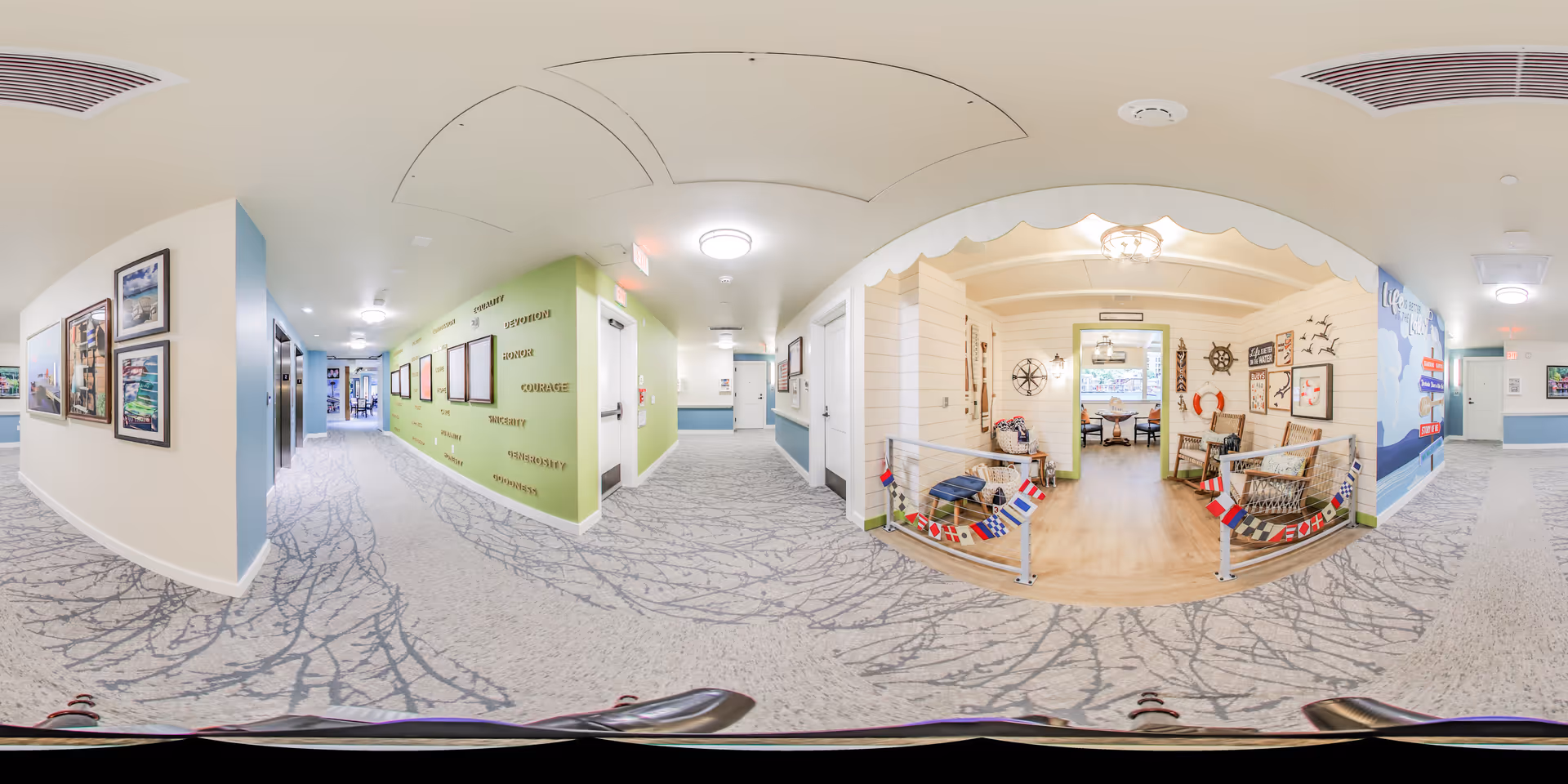 A wide panoramic view of a bright and clean hallway in a senior living facility. The hallway features light-colored walls with framed pictures and motivational words on a green accent wall. To the right, there is a cozy seating area decorated with nautical-themed items such as ship wheels, flags, and life preservers. The floor is carpeted with a tree-branch pattern, and the ceiling has modern light fixtures. The space appears welcoming and well-maintained.