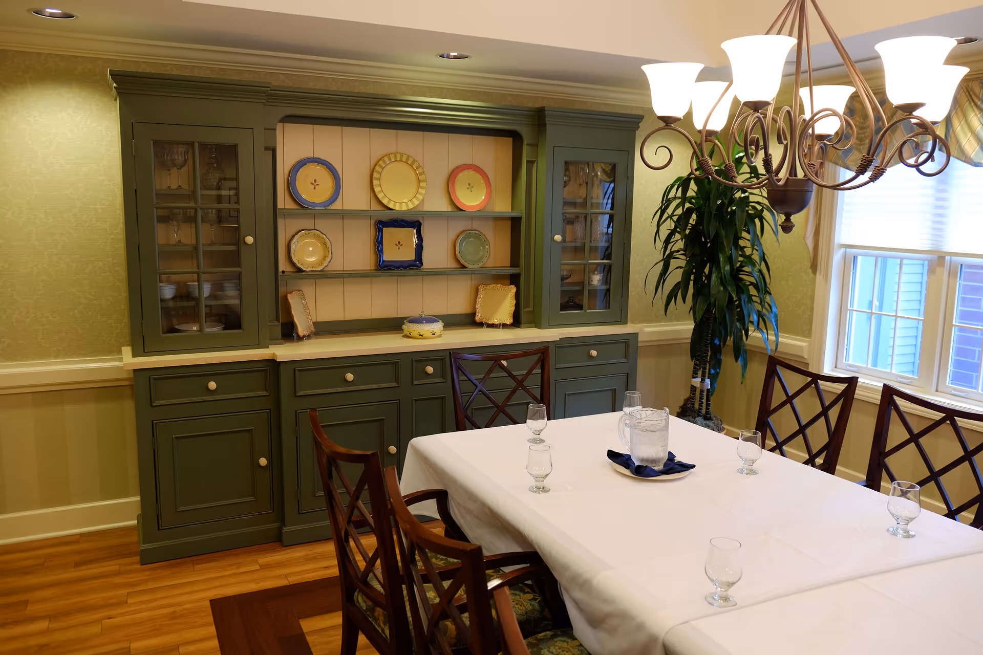 A dining room with a large table covered with a white tablecloth, set with six empty water glasses and a pitcher on a blue napkin. Behind the table is a green wooden cabinet with shelves displaying decorative plates. A large window with blinds and a potted plant are visible to the right, and a chandelier with multiple lights hangs above the table.