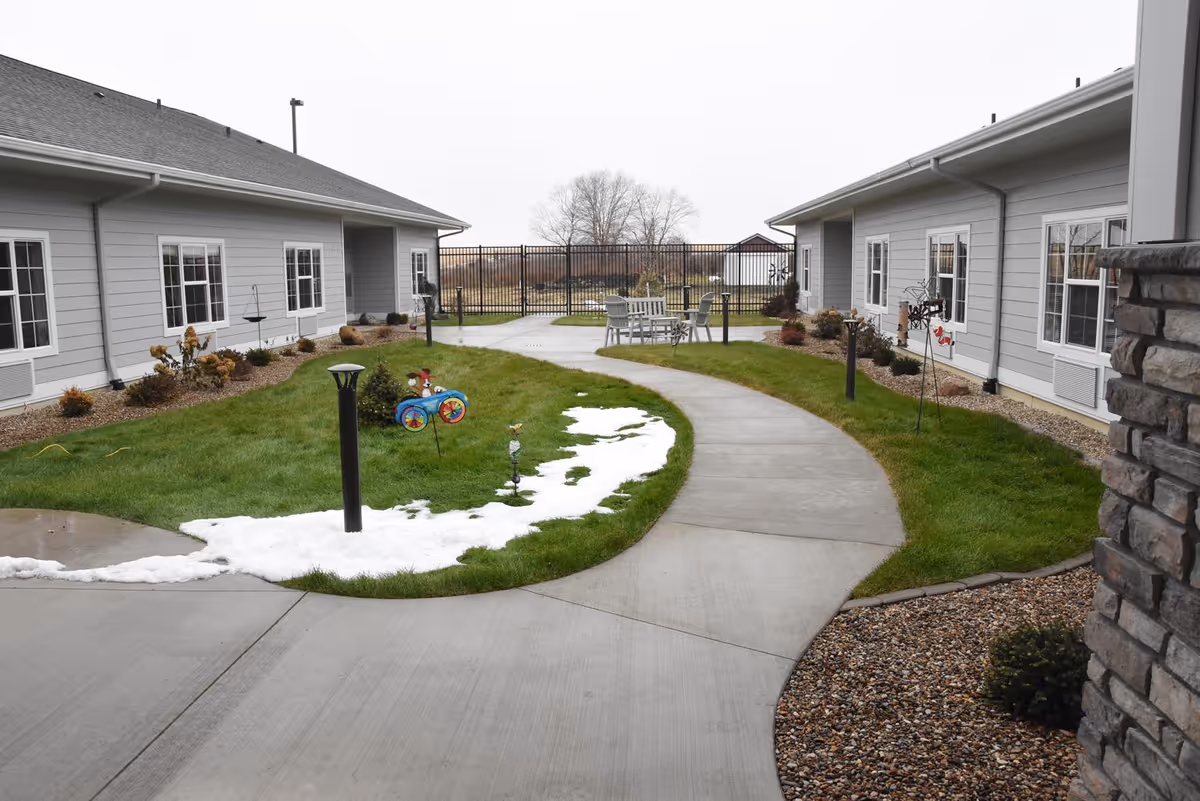 Outdoor courtyard area at Stoney Point Meadows with a curved concrete walkway, green grass patches with some snow, small garden decorations, and two single-story buildings with gray siding and white-framed windows on either side. There is a black metal fence in the background and a seating area with chairs and a table near the fence.