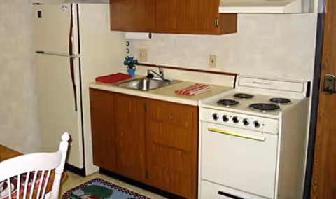 A small kitchen area with a white refrigerator, wooden cabinets, a stainless steel sink, and a white electric stove with four burners. A red towel and a striped cloth are placed on the countertop. Part of a white chair and a colorful rug are visible in the foreground.