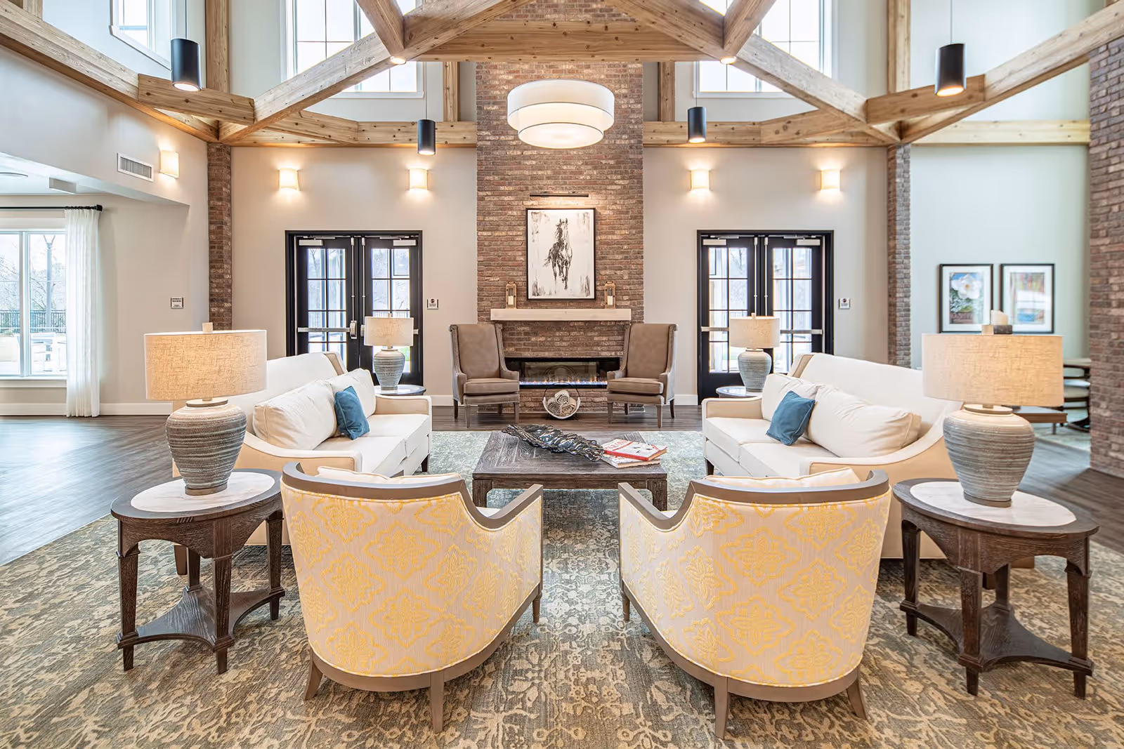 A spacious and well-lit senior living facility lounge area with high wooden beam ceilings, a brick fireplace in the center, two beige sofas with blue accent pillows facing each other, two patterned armchairs in the foreground, wooden side tables with lamps, and large windows and glass doors letting in natural light.