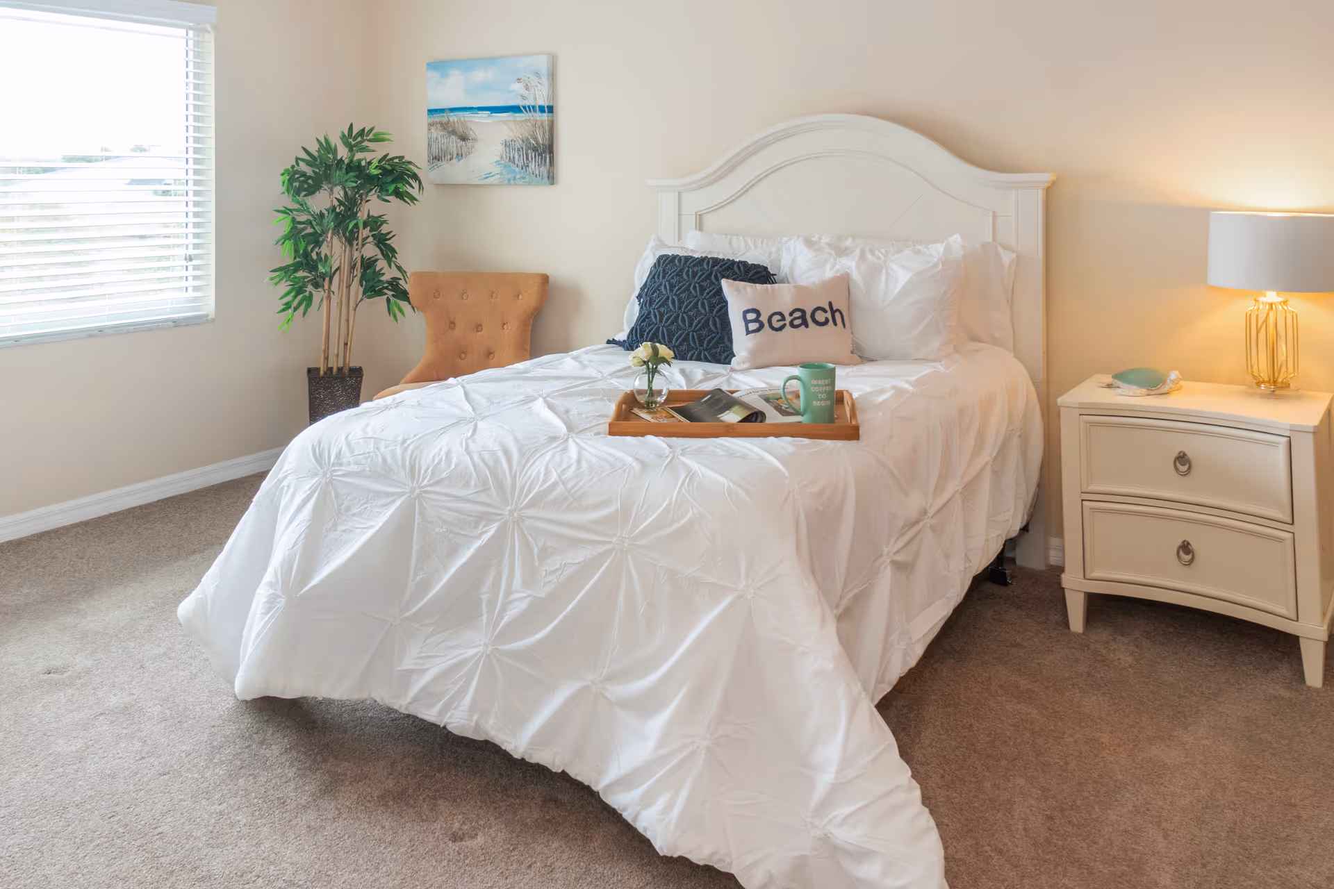 A cozy bedroom with a white bed featuring a headboard, white bedding, and decorative pillows including one that says 'Beach'. There is a wooden tray on the bed with a flower vase, a book, and a green mug. Next to the bed is a cream-colored nightstand with a lamp and a small decorative item. A tan upholstered chair and a potted plant are placed near a window with blinds. A beach-themed painting hangs on the wall above the chair.