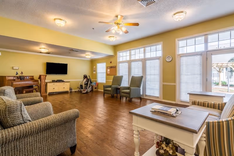 A spacious, well-lit living room in an assisted living facility with wooden flooring, multiple armchairs, a piano, a wall-mounted flat-screen TV, and large windows with blinds allowing natural light to enter. There is also an exercise machine near the back wall and a small table with books and decorative items in the foreground.