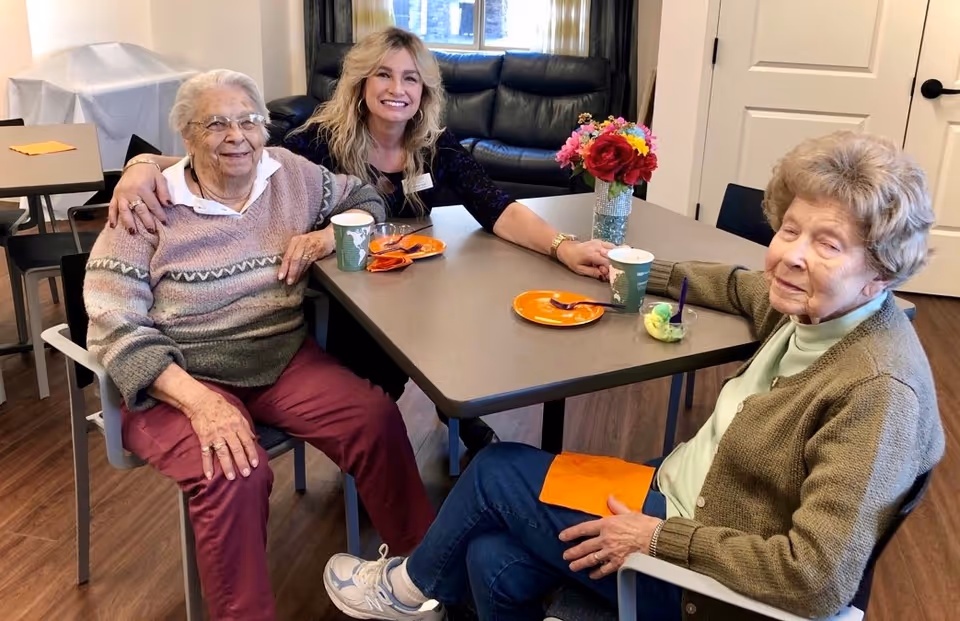 A staff member and two elderly residents sit smiling around a table with cups, plates, and a vase of flowers in a communal dining area.