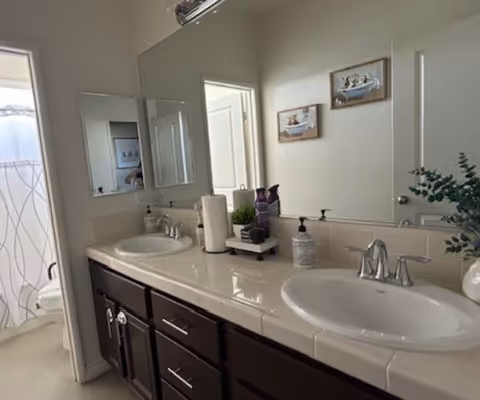 A bathroom with a double sink vanity featuring white tiled countertops and dark wooden cabinets. Above the sinks is a large mirror with two smaller mirrors on the adjacent wall. On the countertop are various items including a paper towel roll, soap dispenser, and decorative plants. In the background, a shower with a white curtain is visible.