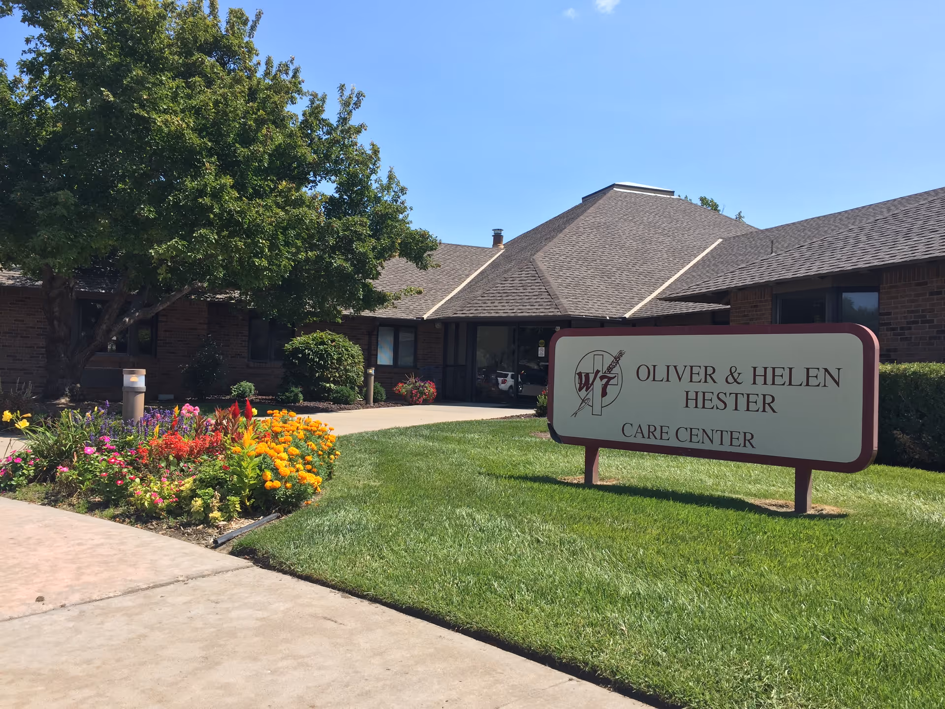 Exterior view of the Oliver & Helen Hester Care Center building with a well-maintained lawn, colorful flower beds, and a large tree under a clear blue sky.