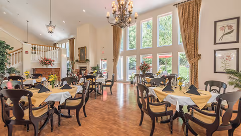 Bright and elegant dining room with multiple round tables covered in white and yellow tablecloths, each set with black napkins and glassware. The room features large floor-to-ceiling windows with brown curtains, a chandelier, wooden flooring, and floral arrangements on tables and near the windows. A staircase and a fireplace are visible in the background.