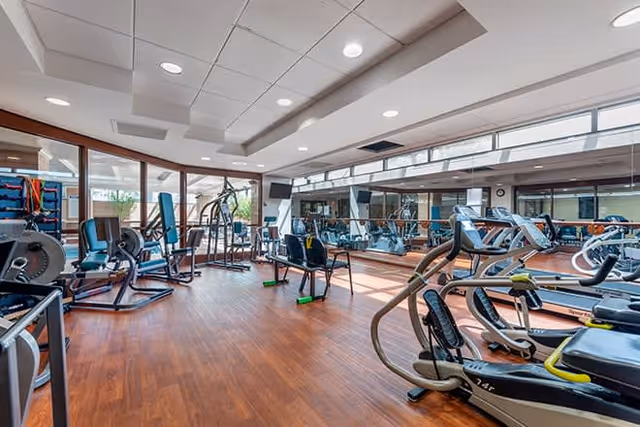 A spacious fitness room with various exercise equipment including treadmills, stationary bikes, weight machines, and chairs. The room has wooden flooring, large windows allowing natural light, and mirrors on one wall.