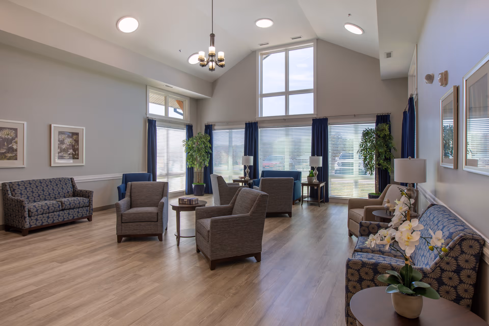 A spacious and well-lit living room area with multiple armchairs and sofas arranged around small tables. The room features large windows with blue curtains, allowing natural light to fill the space. There are framed pictures on the walls, potted plants, and a chandelier hanging from the high ceiling. The floor is wooden, and the overall decor is modern and inviting.
