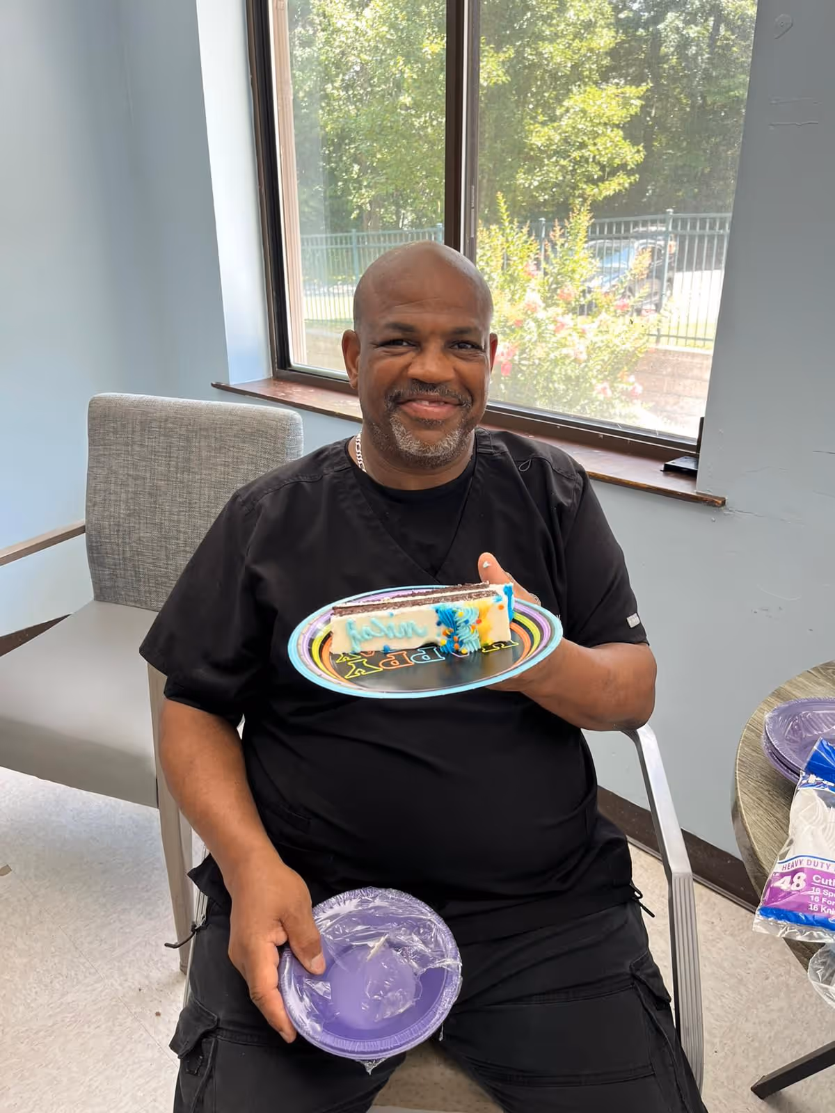 A man wearing black scrubs is sitting on a chair indoors near a window, smiling and holding a colorful plate with a piece of cake and a blue candle shaped like the number 3. He also holds a stack of purple disposable plates wrapped in plastic. There is a table nearby with more disposable plates and utensils.