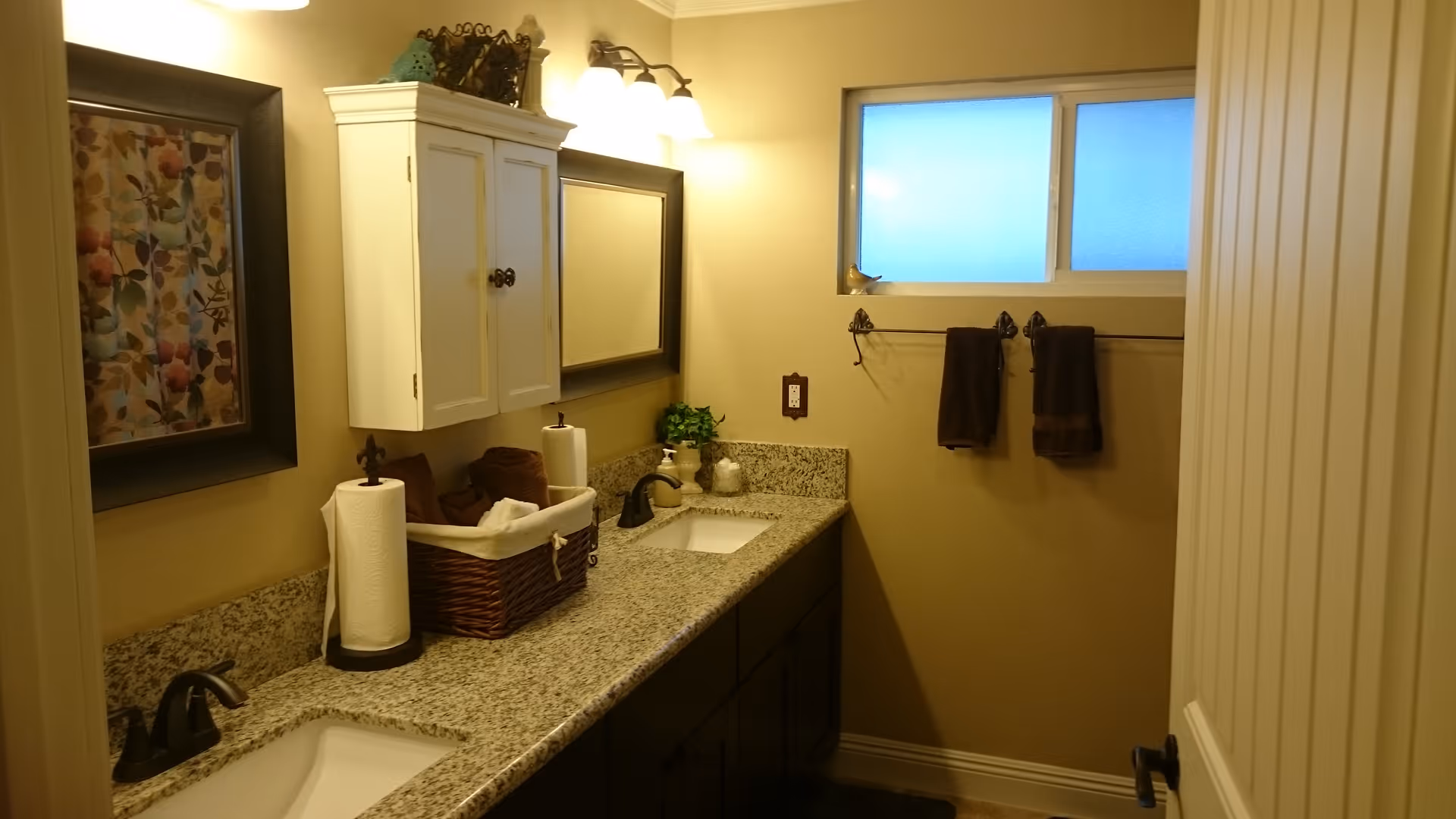 A bathroom with a granite countertop featuring two sinks and dark faucets. Above the countertop is a white cabinet and a rectangular mirror with a light fixture above it. On the countertop, there is a basket with towels, a paper towel holder, a soap dispenser, and a small plant. A framed floral picture hangs on the wall to the left. On the right wall, there is a frosted window and two dark brown towels hanging on a towel rack.