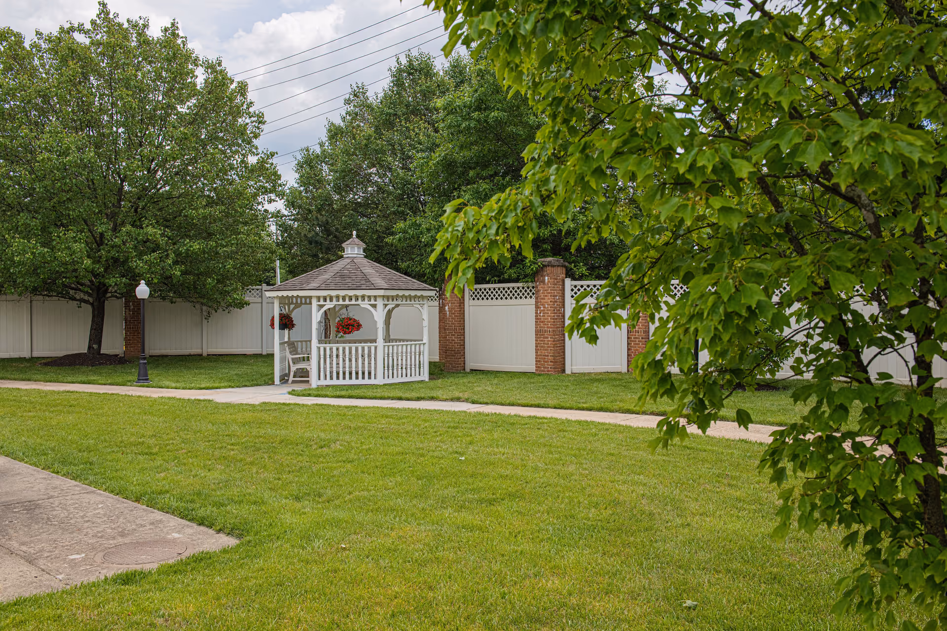 A peaceful outdoor garden area with a white gazebo decorated with hanging red flower baskets. The gazebo is surrounded by green grass, trees, and a white fence with brick pillars. A paved walkway runs through the grassy area.