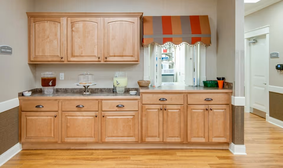 A service counter area in a senior living facility with wooden cabinets and drawers, two beverage dispensers filled with iced tea and lemonade, a covered cake stand with pastries, and a small window with a striped orange and gray awning. The floor is wooden, and there are signs on the walls indicating different rooms.