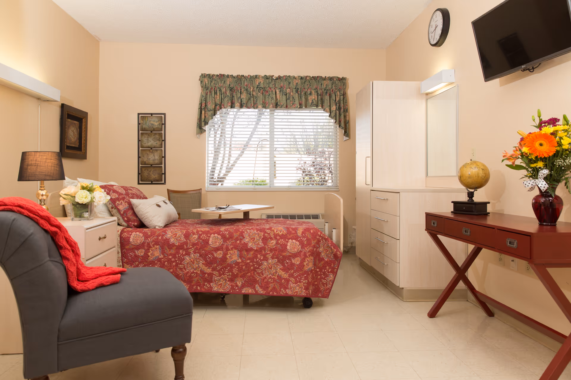 A cozy bedroom in a senior living facility featuring a bed with a red floral bedspread, a small bedside table with a lamp and flowers, a gray upholstered chair with a red throw blanket, a window with floral valance curtains, a white cabinet with drawers, a red wooden desk with a globe and a vase of colorful flowers, a wall-mounted TV, and a clock above the TV.