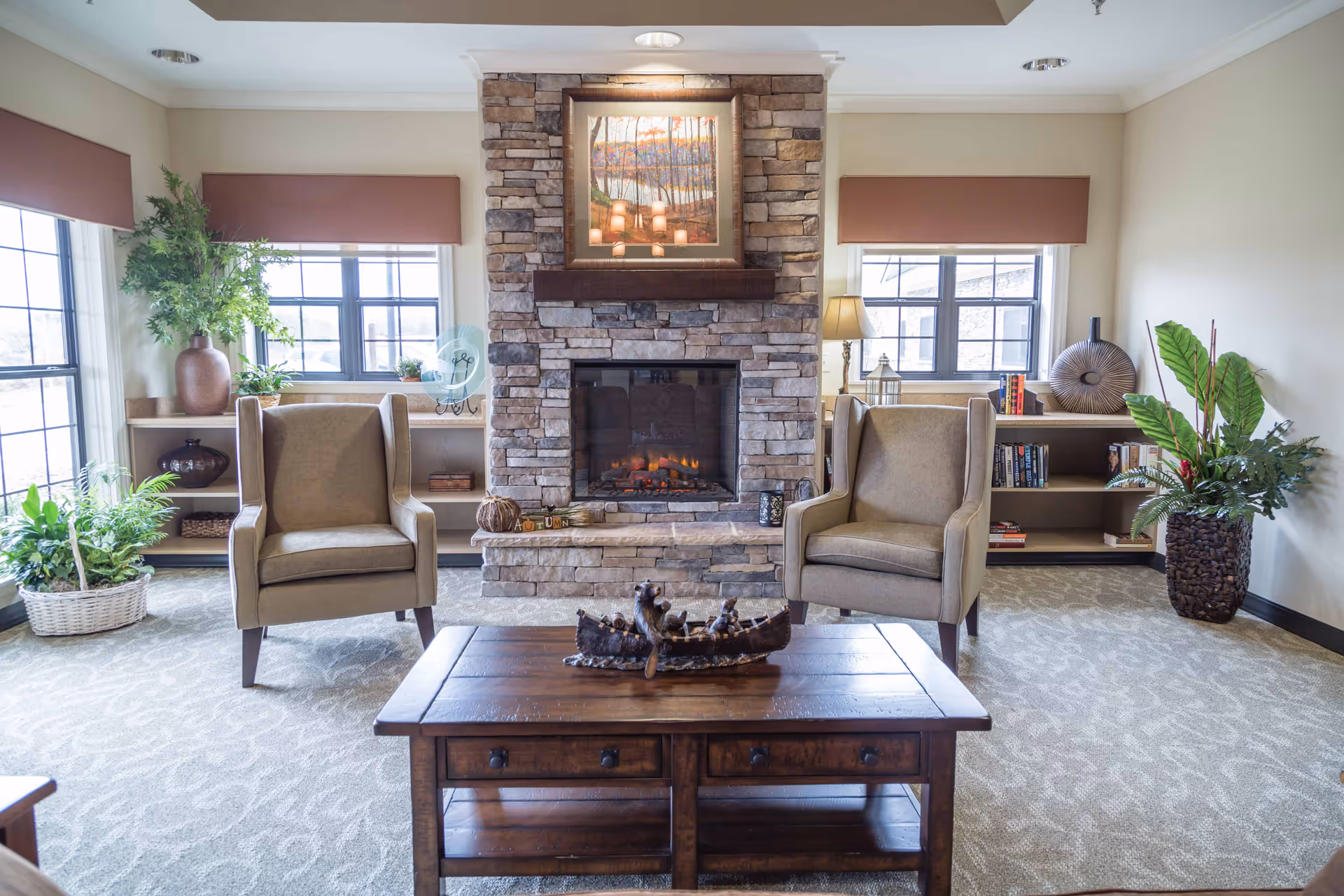 A cozy living room area with two beige armchairs facing a stone fireplace. Above the fireplace is a framed painting of a forest scene. There are two windows with brown valances on either side of the fireplace. Bookshelves with decorative items and books flank the fireplace. A wooden coffee table with a decorative centerpiece is in the foreground, and there are several green plants placed around the room.