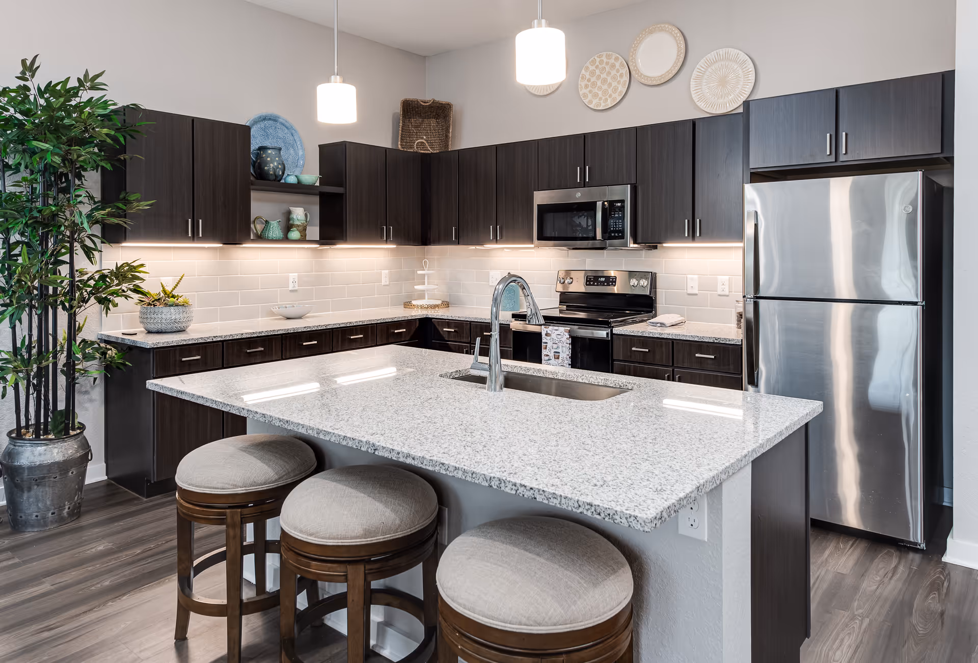 Modern kitchen with a granite island and three round stools, dark cabinets, stainless steel appliances, pendant lights, and decorative accents.