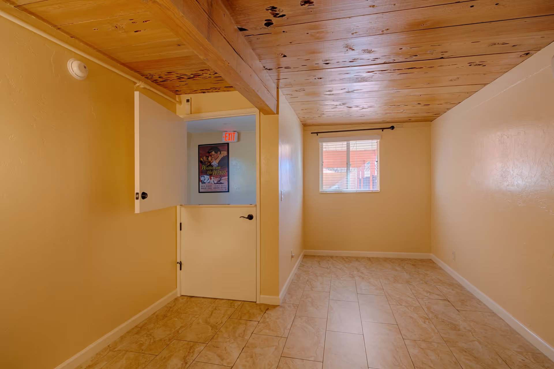 Empty narrow room with tiled floor, wood-plank ceiling, a small window with blinds, and a Dutch-style half door opening to a hallway.