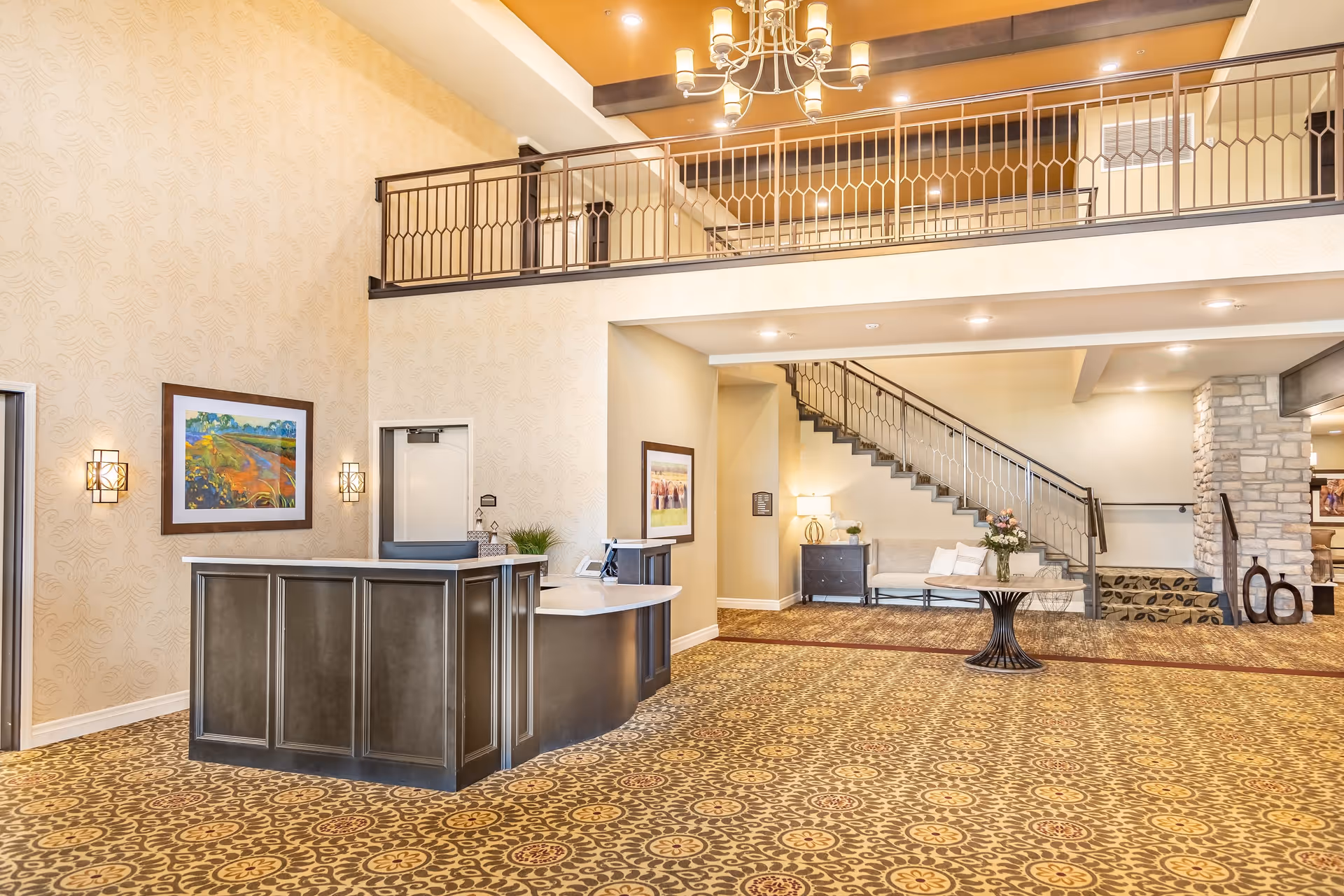 Spacious and well-lit lobby area with a reception desk, patterned carpet, a staircase with metal railing, a round table with a flower arrangement, framed artwork on the walls, and a chandelier hanging from the ceiling.