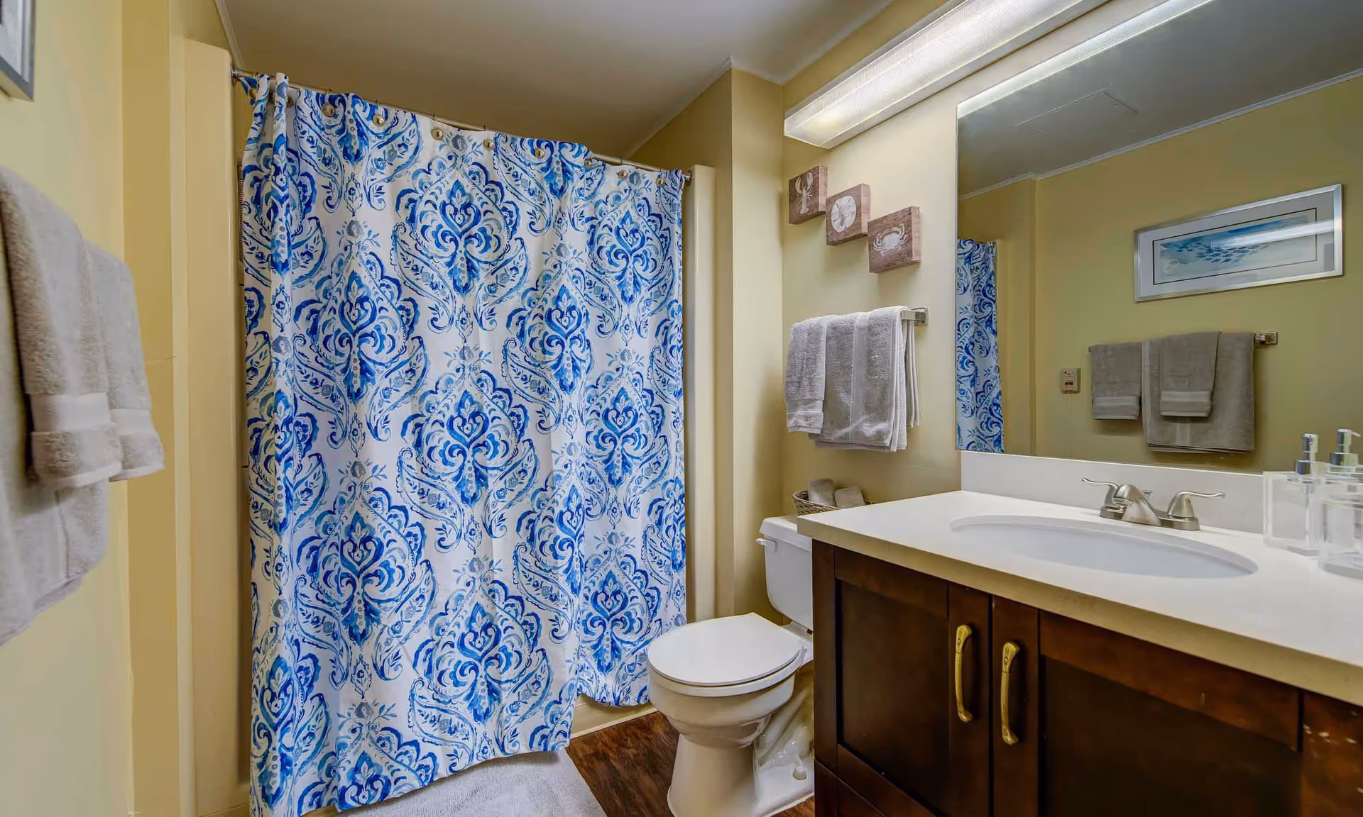 A bathroom with a white toilet, a wooden vanity with a white countertop and sink, a large mirror above the sink, and a shower with a blue and white patterned curtain. There are white towels hanging on racks and three decorative wall plaques above the toilet.